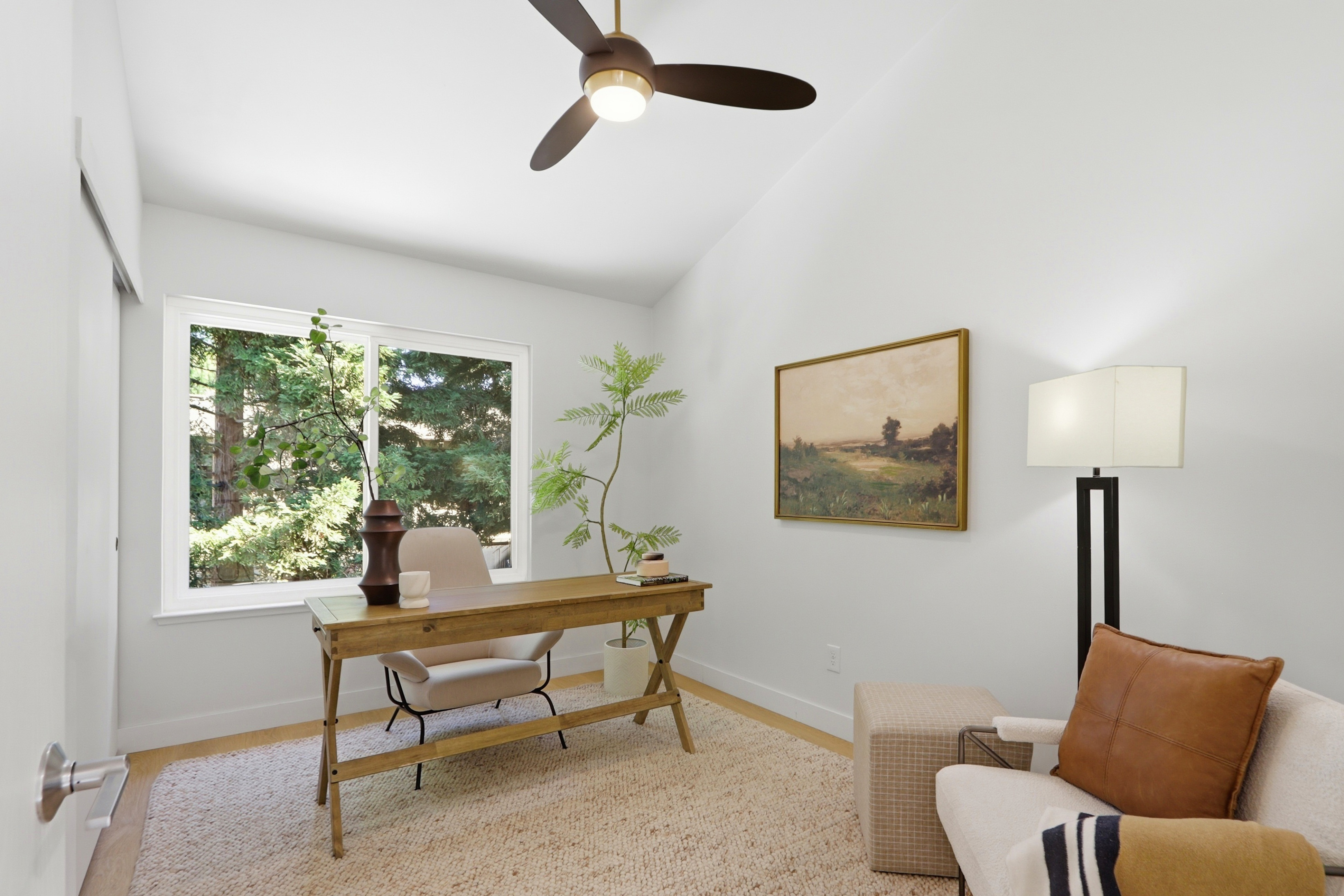 This bright and airy home office features a minimalist aesthetic with a wooden desk, a comfortable cream-colored chair, and a cozy reading nook in the corner. A large window provides ample natural light and a view of lush greenery, while a ceiling fan and a floor lamp offer functional lighting. The space is accented by a textured area rug, a potted plant, and a landscape painting, creating a serene and productive atmosphere.