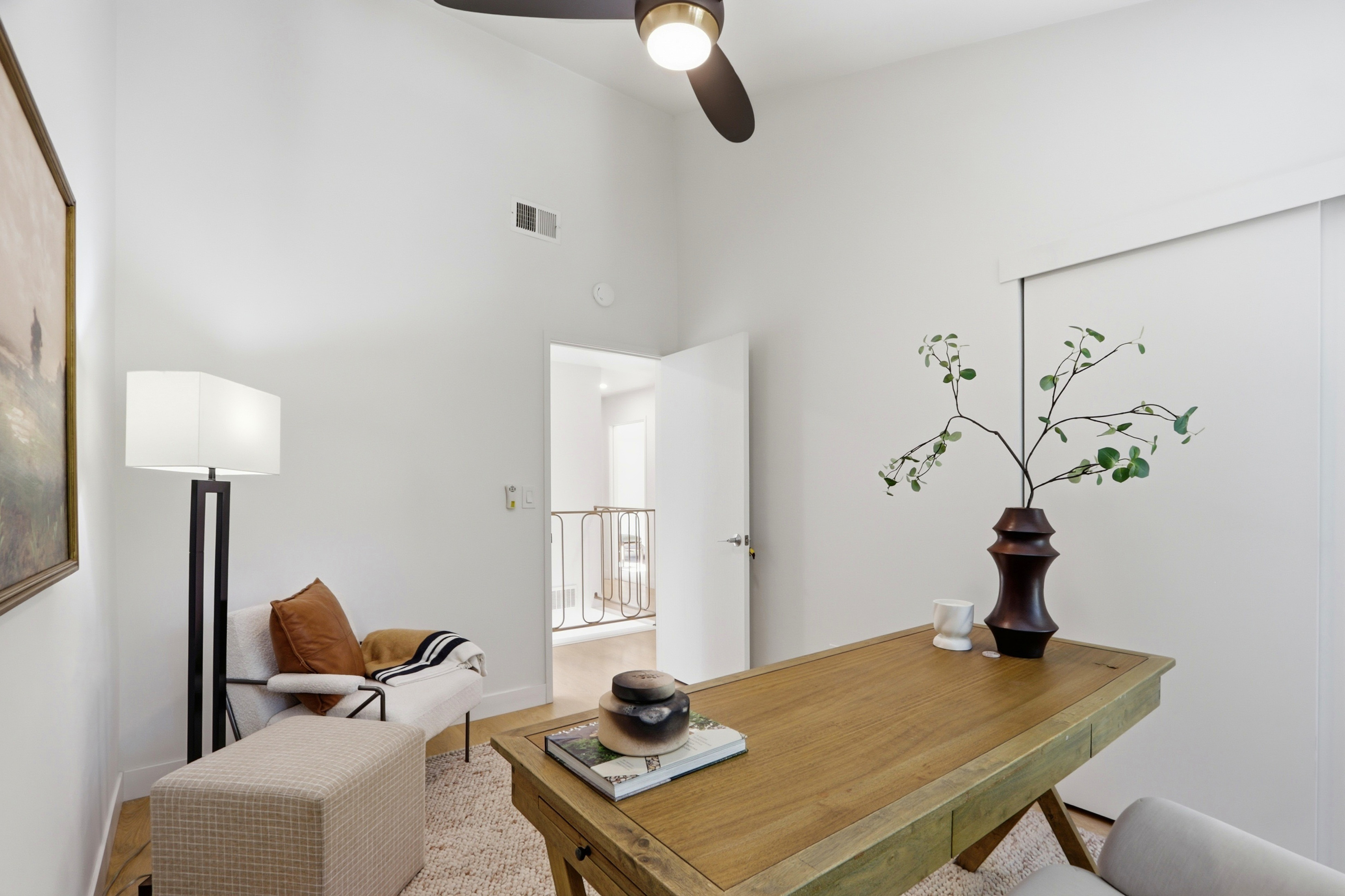 This bright and airy home office features a wooden desk as the centerpiece, complemented by a cozy reading nook with a plush armchair and ottoman. The room is accented with minimalist decor, including a vase with greenery and a framed piece of art, all set against clean white walls and high ceilings. The perspective captures the inviting workspace, looking toward an open doorway that reveals a glimpse of a hallway with a decorative railing.