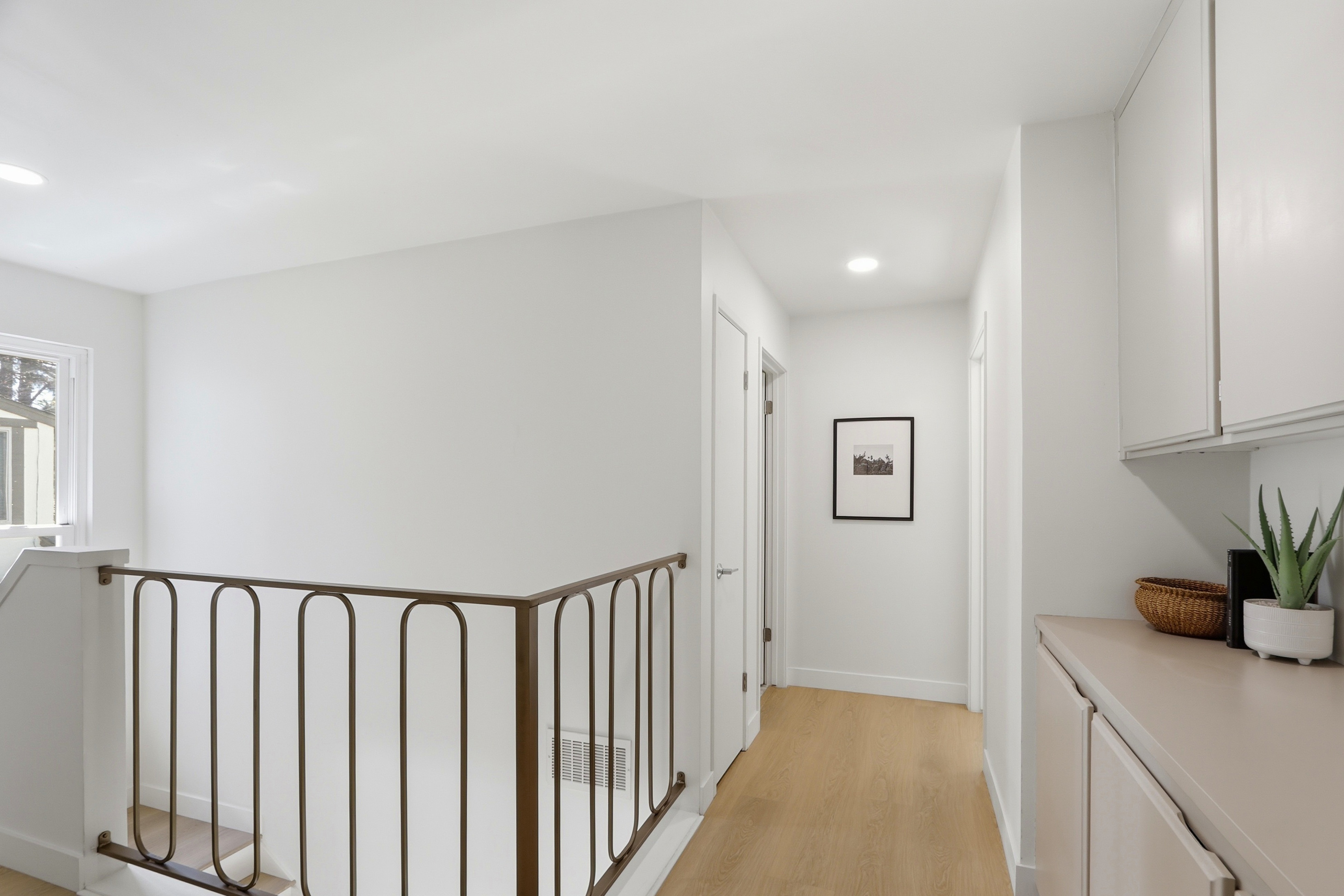 This bright and minimalist hallway features light wood-tone flooring and crisp white walls, creating an airy and welcoming atmosphere. A distinctive bronze-toned metal railing lines the staircase, while a framed piece of art adds a touch of sophistication to the corridor. To the right, a built-in cabinet provides convenient storage, topped with a small potted plant that adds a subtle organic element to the clean, modern aesthetic.