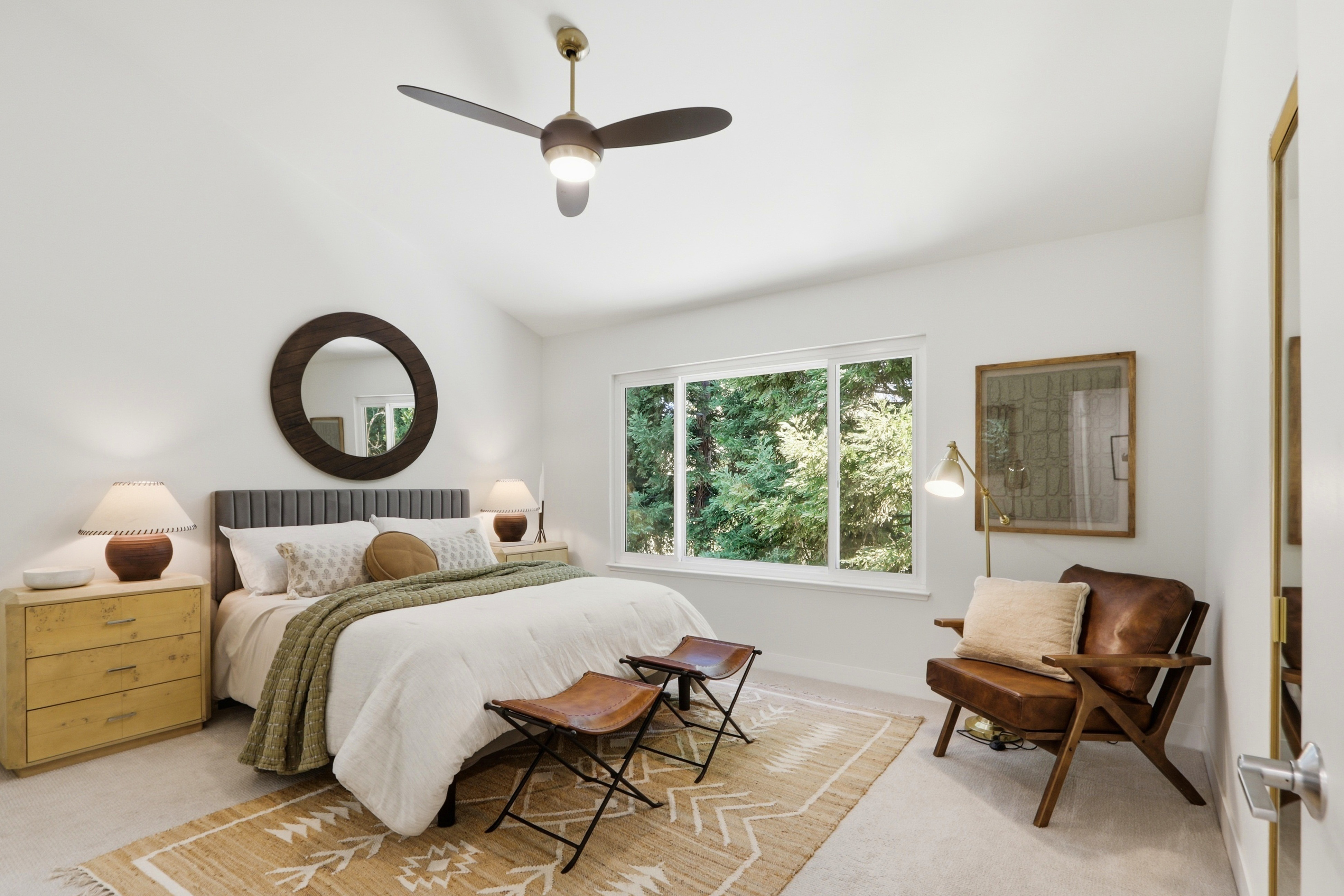 This bright and airy primary bedroom features a neutral color palette with warm wood accents and a large window overlooking lush greenery. The room is furnished with a plush bed, two leather stools, and a mid-century modern leather armchair, creating a cozy and inviting atmosphere. A circular mirror above the headboard and a stylish ceiling fan add contemporary character to the space.