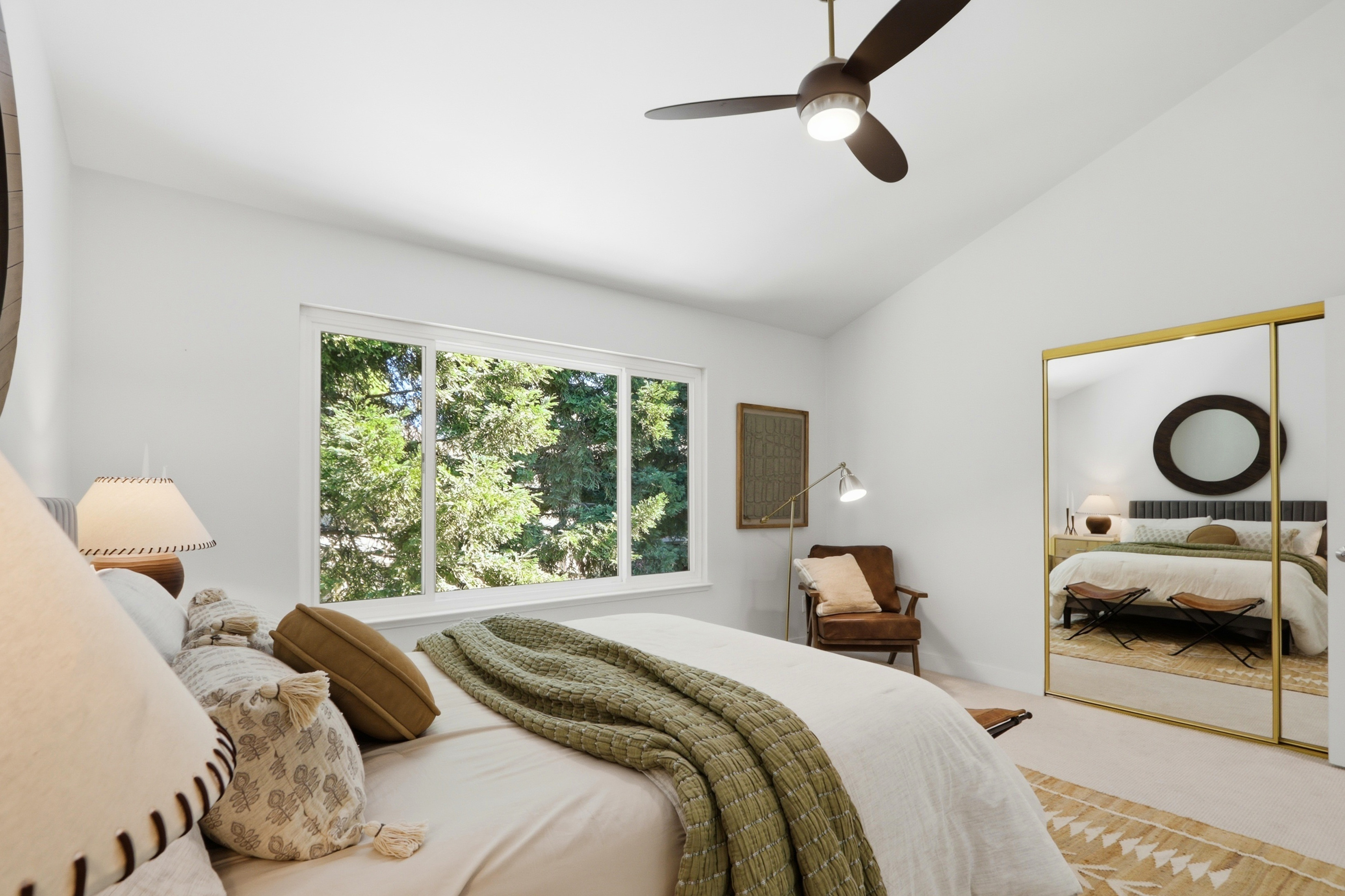 This bright and airy primary bedroom features a neutral color palette with soft green and earthy brown accents, creating a serene and inviting atmosphere. A large window provides ample natural light and a view of lush greenery, while a mirrored closet door adds depth and reflects the room's elegant decor. The space is completed by a modern ceiling fan, a cozy leather reading chair, and a well-appointed bed with layered textiles.