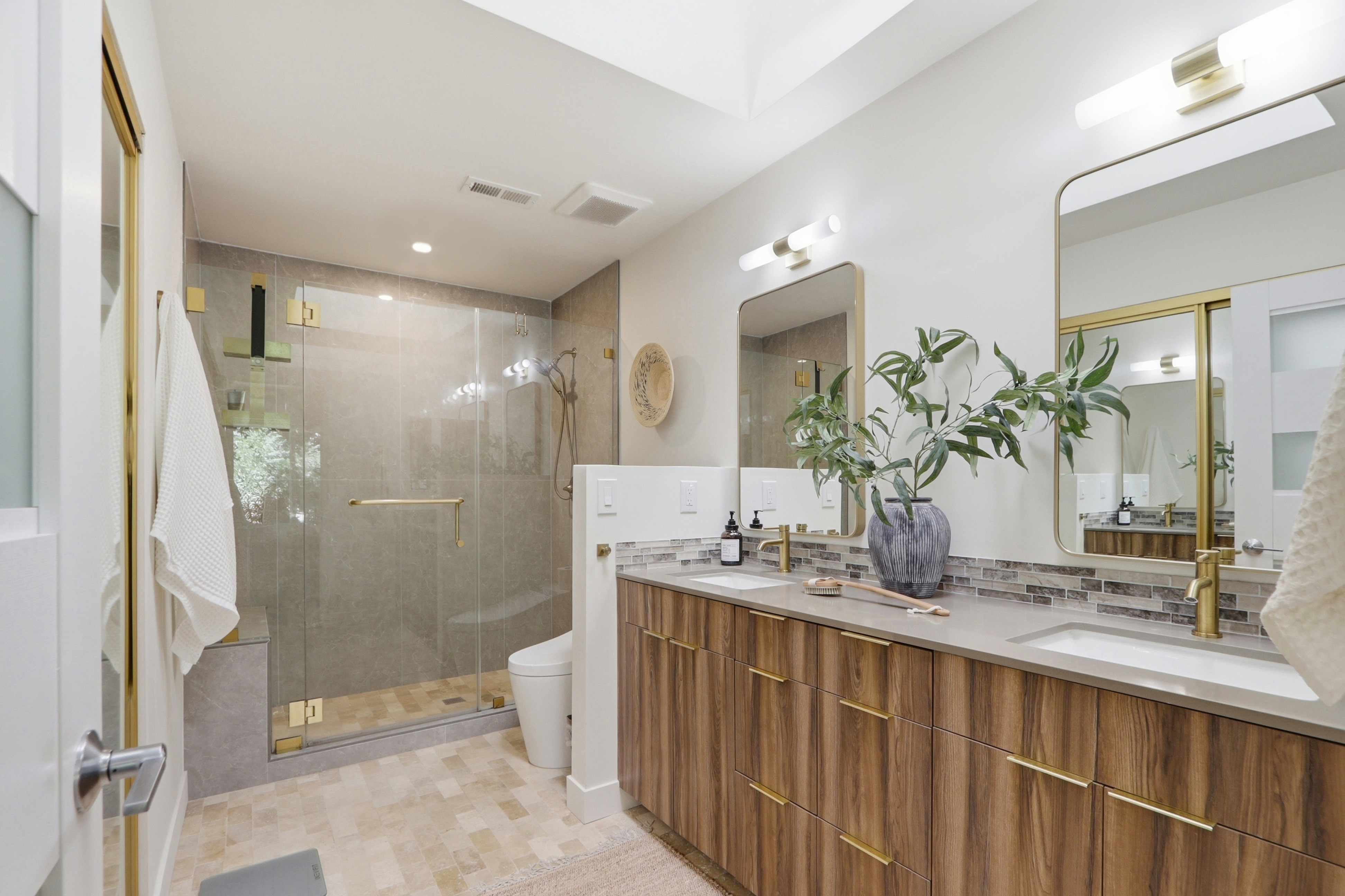This modern primary bathroom features a sleek double vanity with wood-grain cabinetry, gold hardware, and a stone countertop. A spacious walk-in shower with glass doors and neutral tile work sits adjacent to the vanity, while gold-framed mirrors and contemporary lighting fixtures add a touch of elegance. The overall aesthetic is clean, sophisticated, and spa-like, captured from a wide-angle perspective that highlights the functional layout.