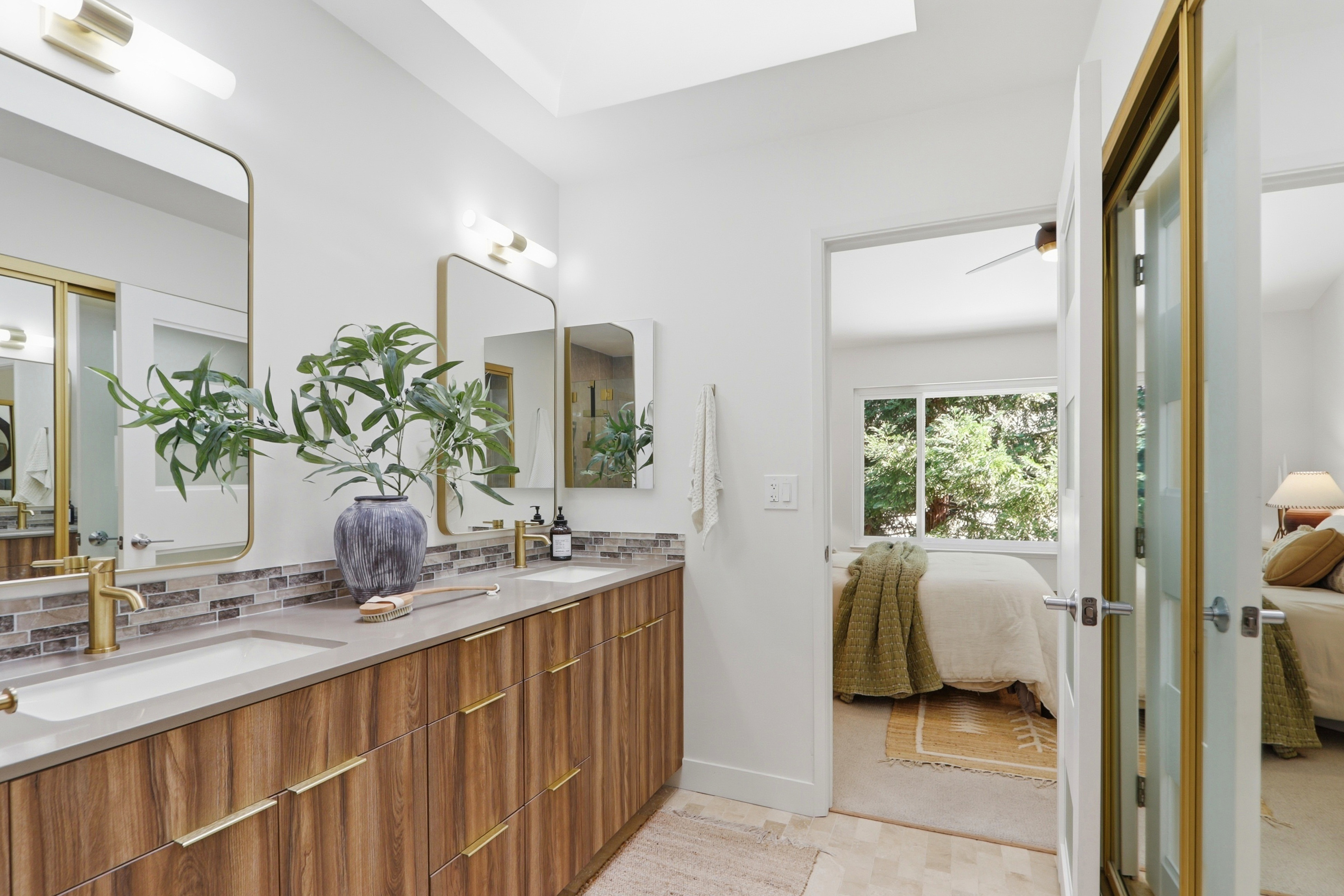 This modern primary bathroom features a sleek dual-sink vanity with wood-grain cabinetry, gold hardware, and a stone countertop. The space is bright and airy, with a seamless transition into the adjacent bedroom, which is visible through the open doorway. The design combines contemporary aesthetics with warm, natural textures, creating a sophisticated and inviting atmosphere.