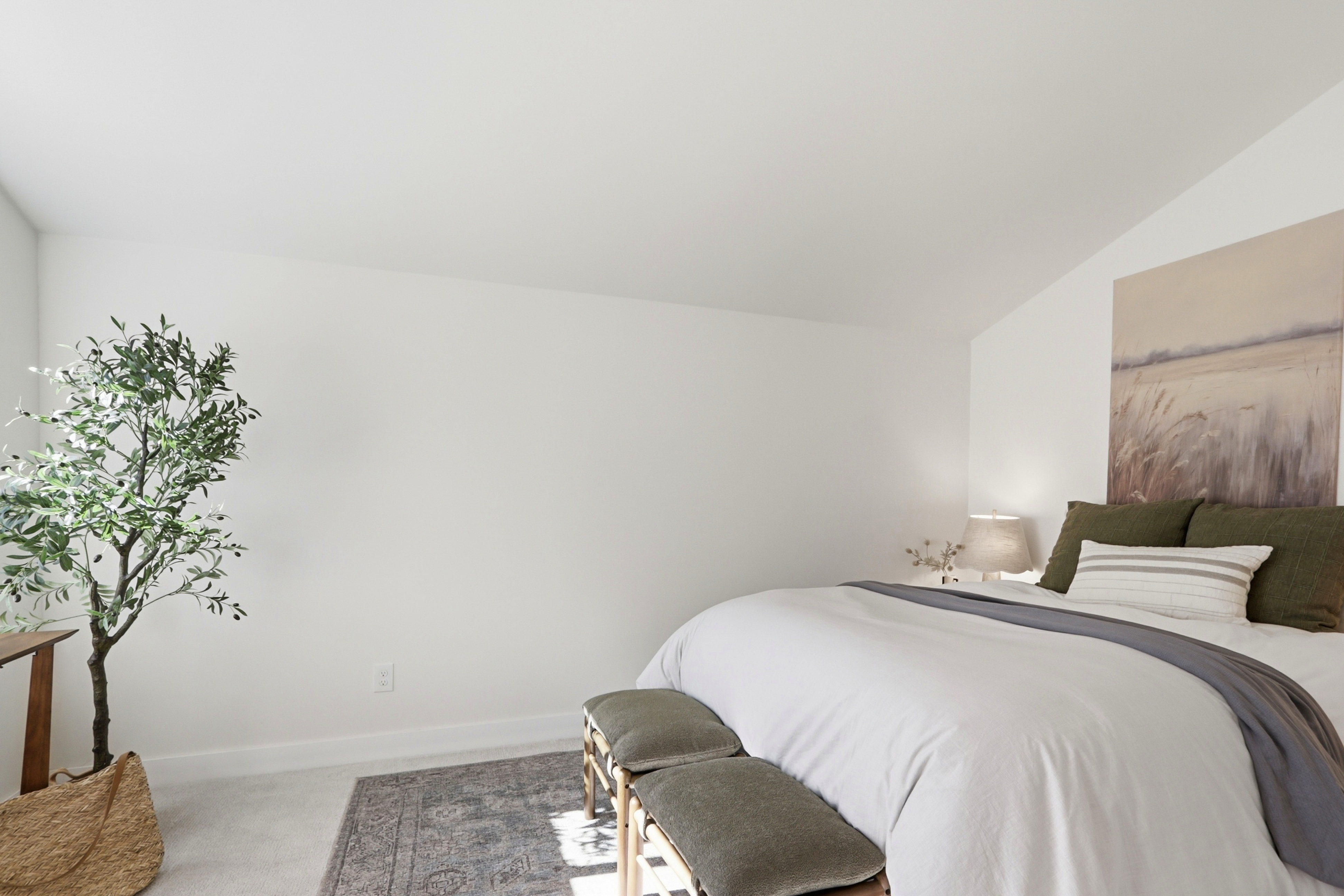 This serene primary bedroom features a neutral color palette with soft white walls and a plush white duvet, accented by olive green pillows and a matching bench at the foot of the bed. A large, calming landscape painting hangs above the headboard, while a potted olive tree in a woven basket adds a touch of organic texture to the corner. The room is bathed in soft, natural light, creating a peaceful and inviting atmosphere.