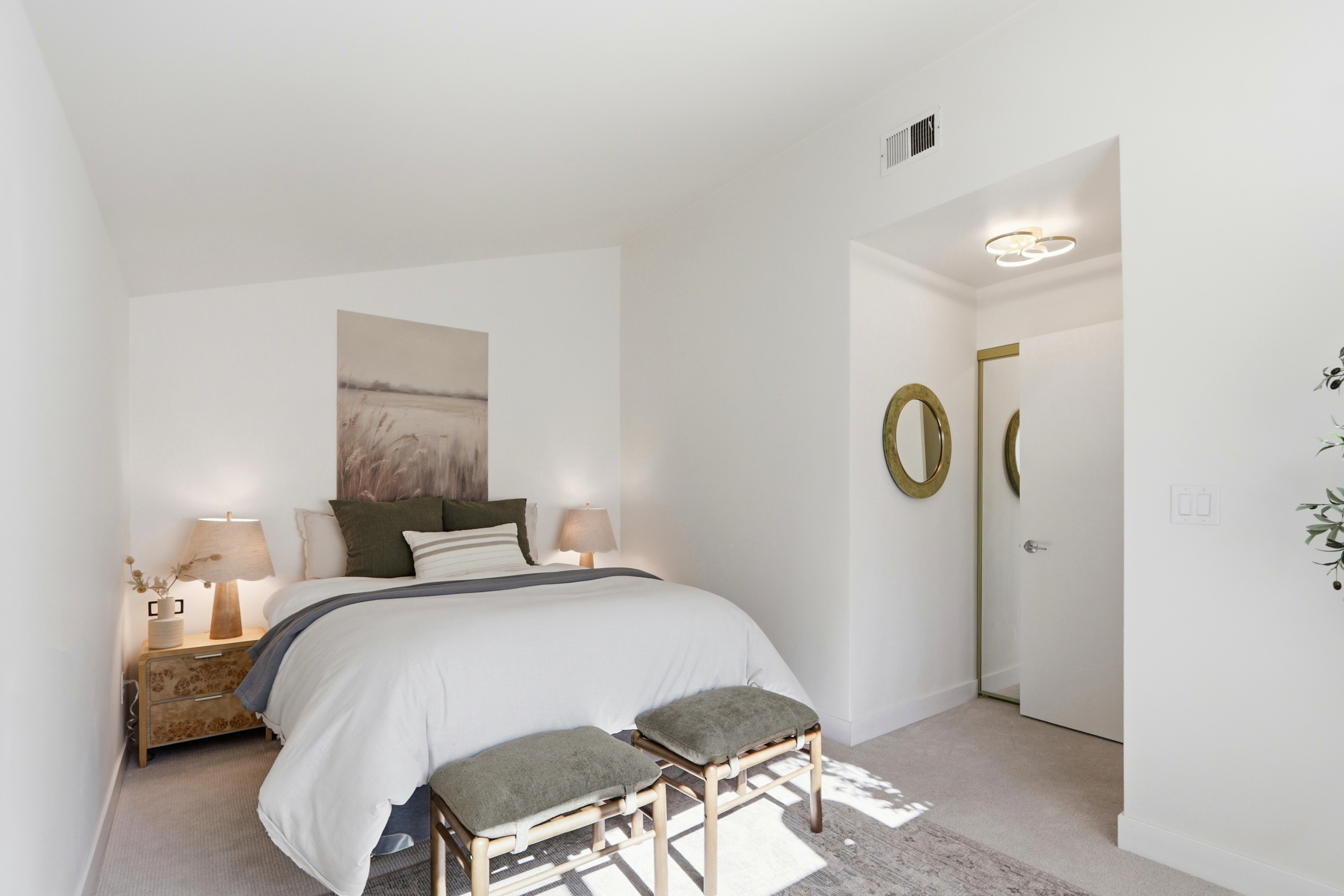 This serene primary bedroom features a neutral color palette with soft textures, centered around a plush bed adorned with olive green pillows and a white duvet. Two wooden stools sit at the foot of the bed, while a bedside table with a textured lamp adds warmth to the space. The room includes a small alcove with a mirror and a modern ceiling light, creating a clean, inviting, and contemporary atmosphere.