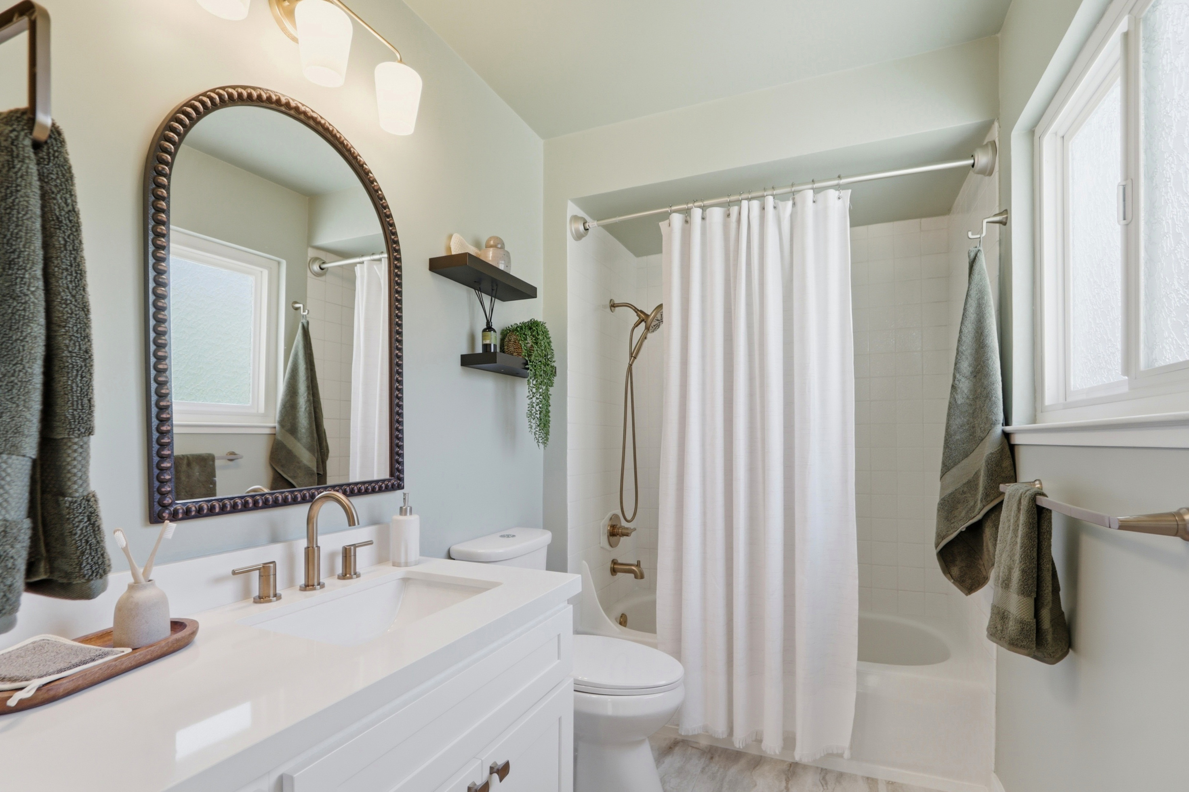 This bright and clean bathroom features a white vanity with a quartz countertop, a stylish arched mirror, and a shower-tub combination with a white curtain. The space is accented with sage green towels and wall paint, creating a calming, modern aesthetic. The perspective is a straight-on shot from the doorway, showcasing the efficient layout and contemporary fixtures.