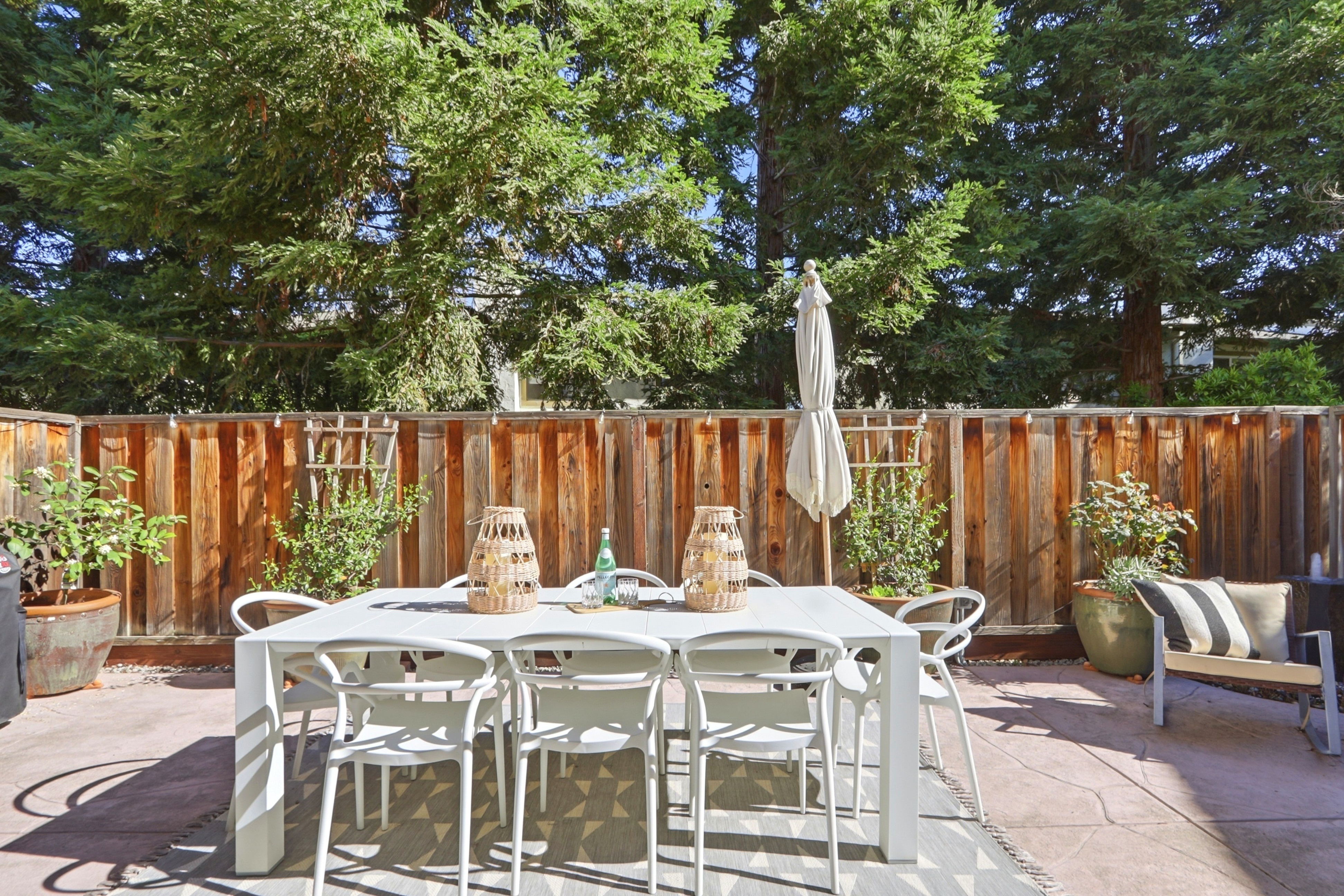 This inviting outdoor patio space features a modern white dining set arranged on a geometric patterned rug, perfect for al fresco entertaining. The area is enclosed by a warm-toned wooden fence and lush greenery, creating a private and serene atmosphere. A closed patio umbrella and potted plants add to the comfortable, well-maintained aesthetic of this backyard retreat.