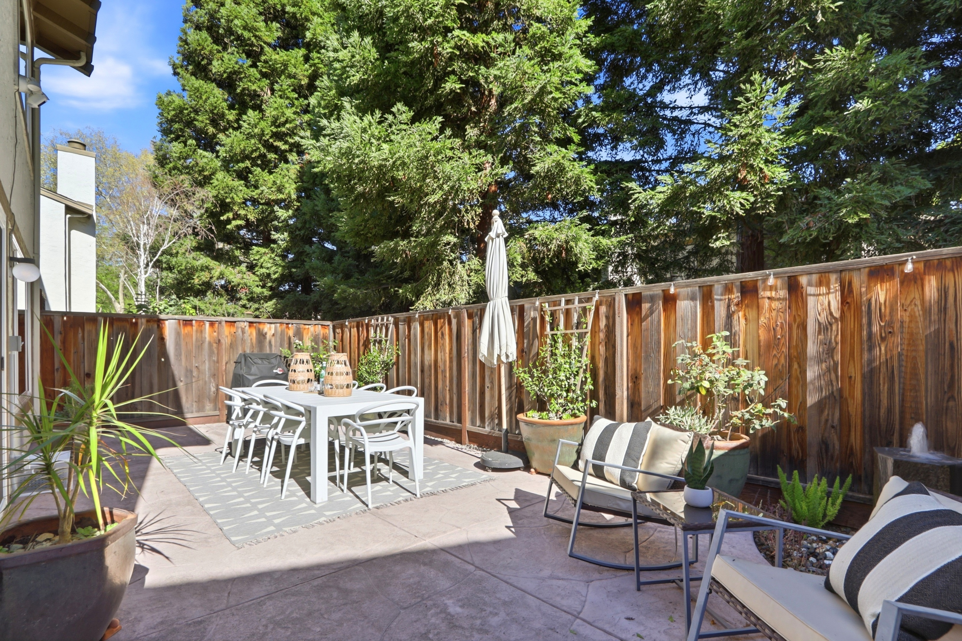 This inviting outdoor patio space features a modern dining set and comfortable lounge chairs, perfect for al fresco entertaining. The area is enclosed by a rustic wood fence and surrounded by lush greenery, creating a private and serene atmosphere. A patterned outdoor rug and potted plants add a touch of style to the concrete patio, which is bathed in warm, natural sunlight.