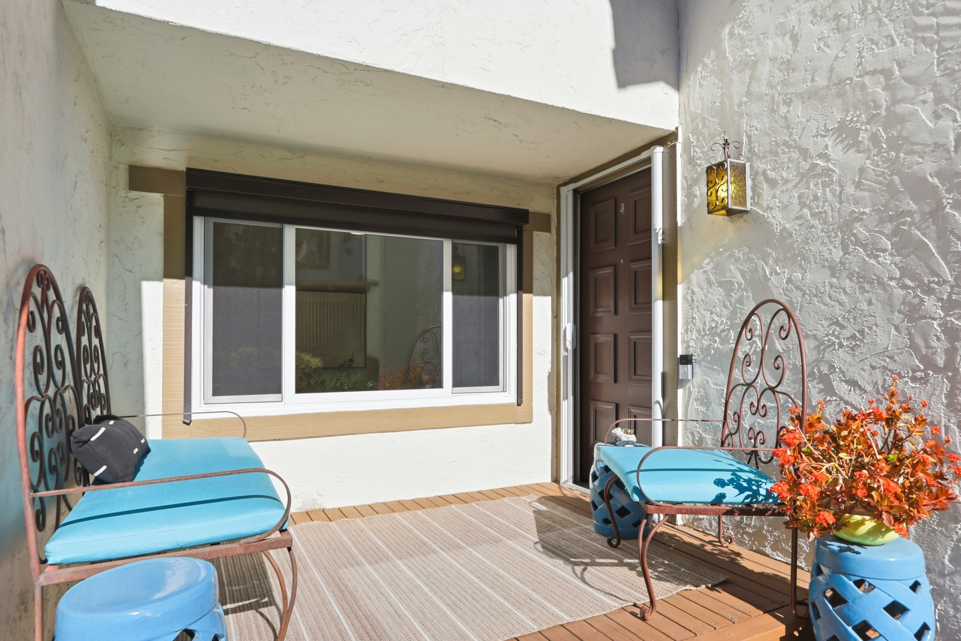 This inviting front porch area features a warm wooden deck furnished with two ornate metal chairs topped with vibrant blue cushions. A large window with a dark frame sits beside the brown front door, while a decorative wall-mounted lantern adds a touch of charm to the textured stucco wall. The space is accented by a potted plant with orange flowers and blue ceramic stools, creating a cozy and welcoming entryway.