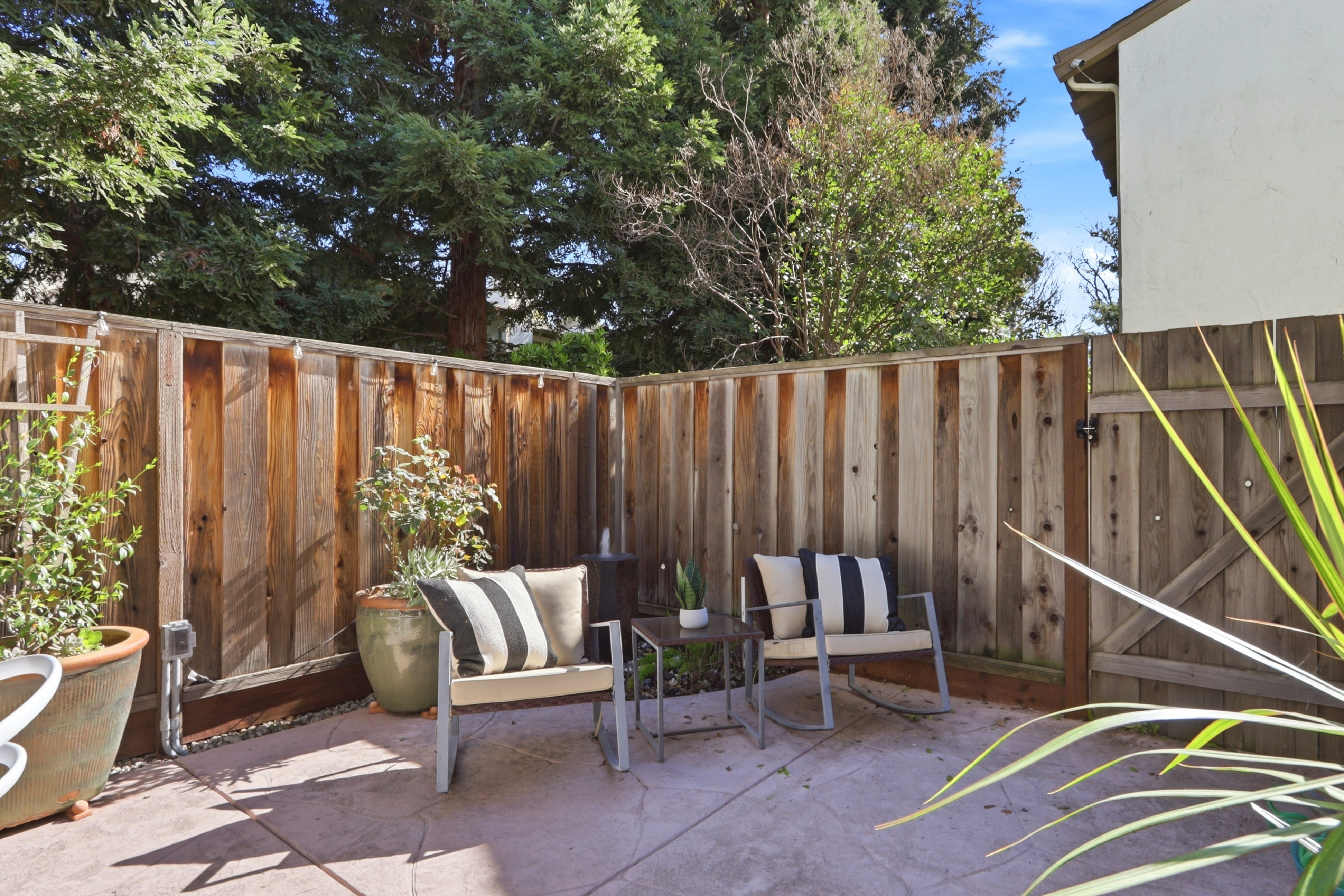 This cozy, private patio space features two comfortable rocking chairs with striped cushions flanking a small side table, creating an inviting outdoor seating area. The space is enclosed by a rustic wooden fence and accented with potted greenery, offering a serene and intimate atmosphere. The warm, natural lighting highlights the textured concrete flooring, making it an ideal spot for relaxation.