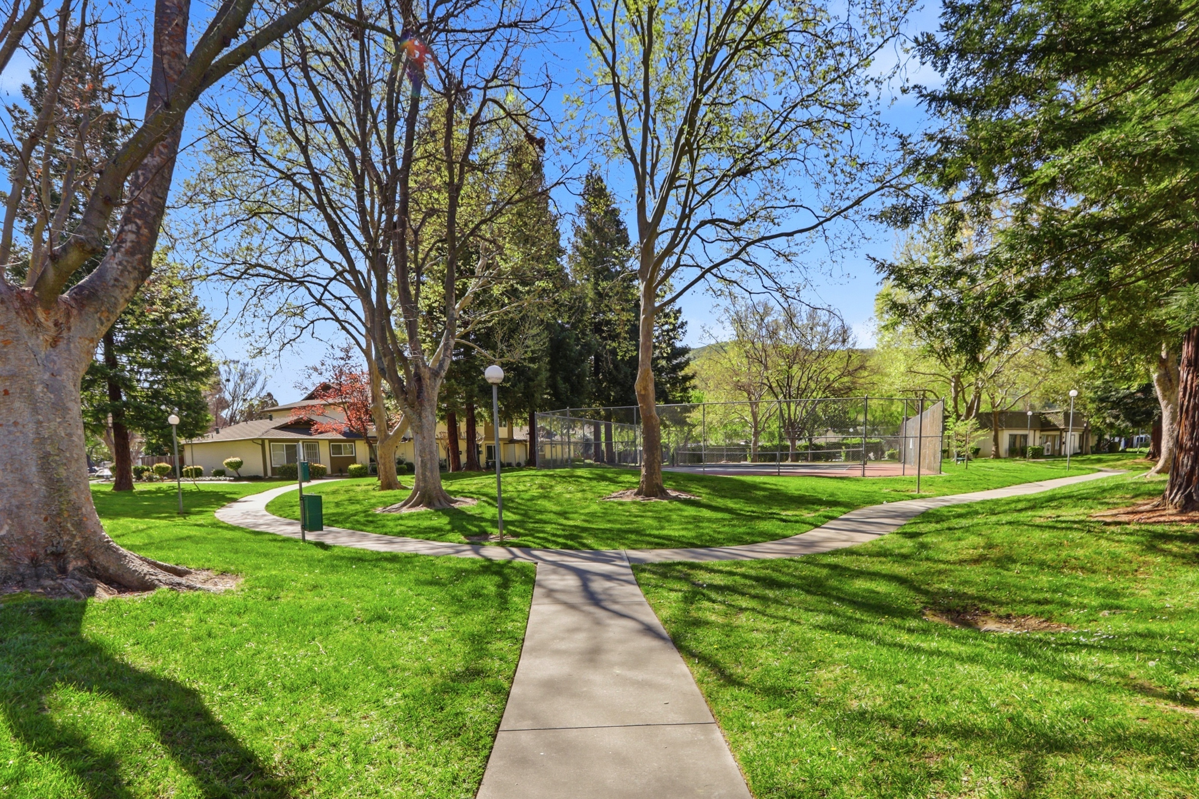 This image captures a serene community park area featuring a well-maintained lawn, mature trees, and a paved walking path that leads toward a fenced tennis court. The scene is bathed in bright, natural sunlight, creating an inviting and peaceful atmosphere for residents. The perspective is a ground-level shot looking down the path, emphasizing the open green space and recreational amenities available within the community.