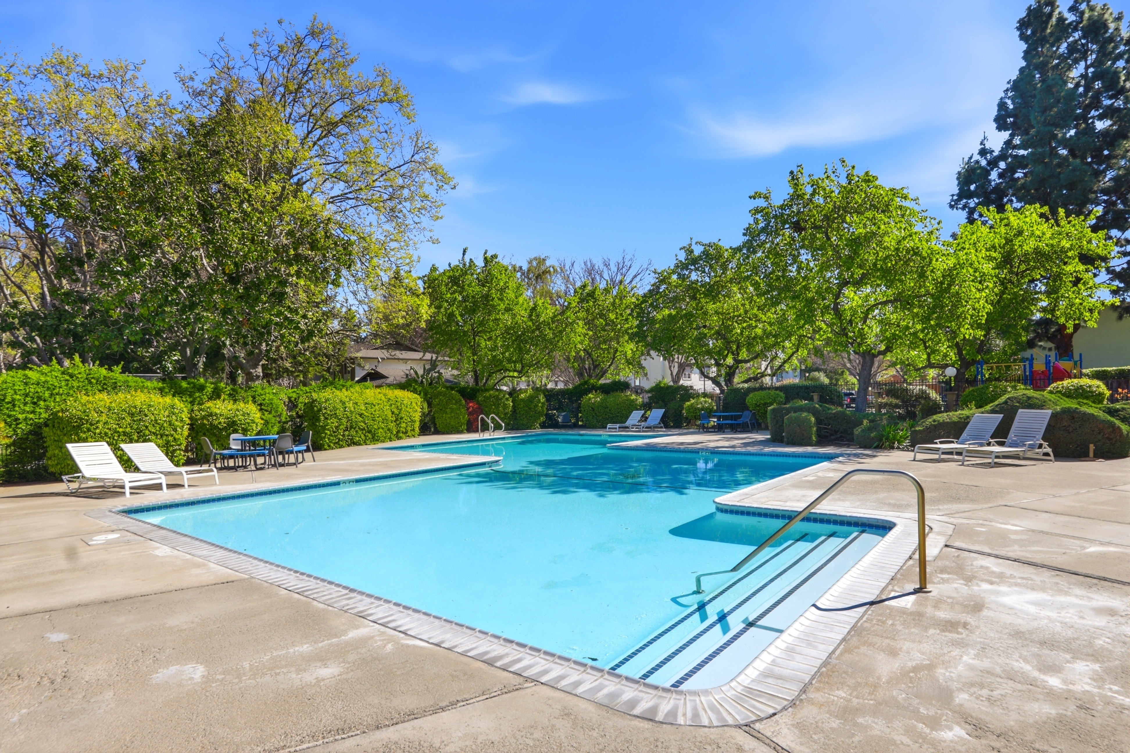 This inviting community swimming pool features a spacious concrete deck surrounded by lush green trees and manicured hedges. The area is furnished with multiple lounge chairs and small dining sets, creating a relaxing atmosphere for residents. The bright, sunny day highlights the clear blue water and the well-maintained landscaping of this outdoor amenity.