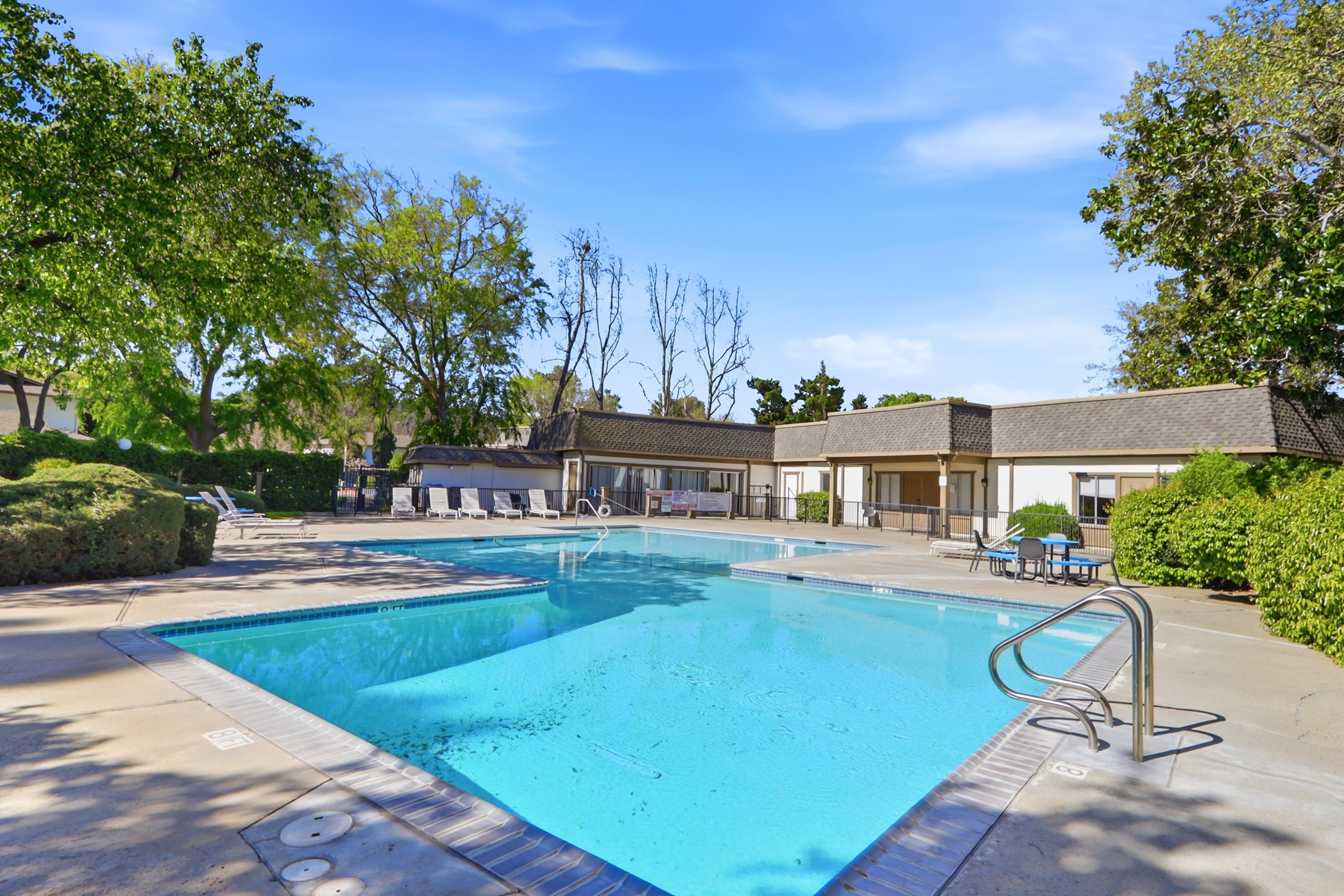 This image showcases a well-maintained community swimming pool set against a clear blue sky, surrounded by lush green trees and manicured landscaping. The pool area features a spacious concrete deck with lounge chairs, a small dining set, and a metal handrail leading into the water. The scene conveys a relaxing, resort-style atmosphere, perfect for community recreation.