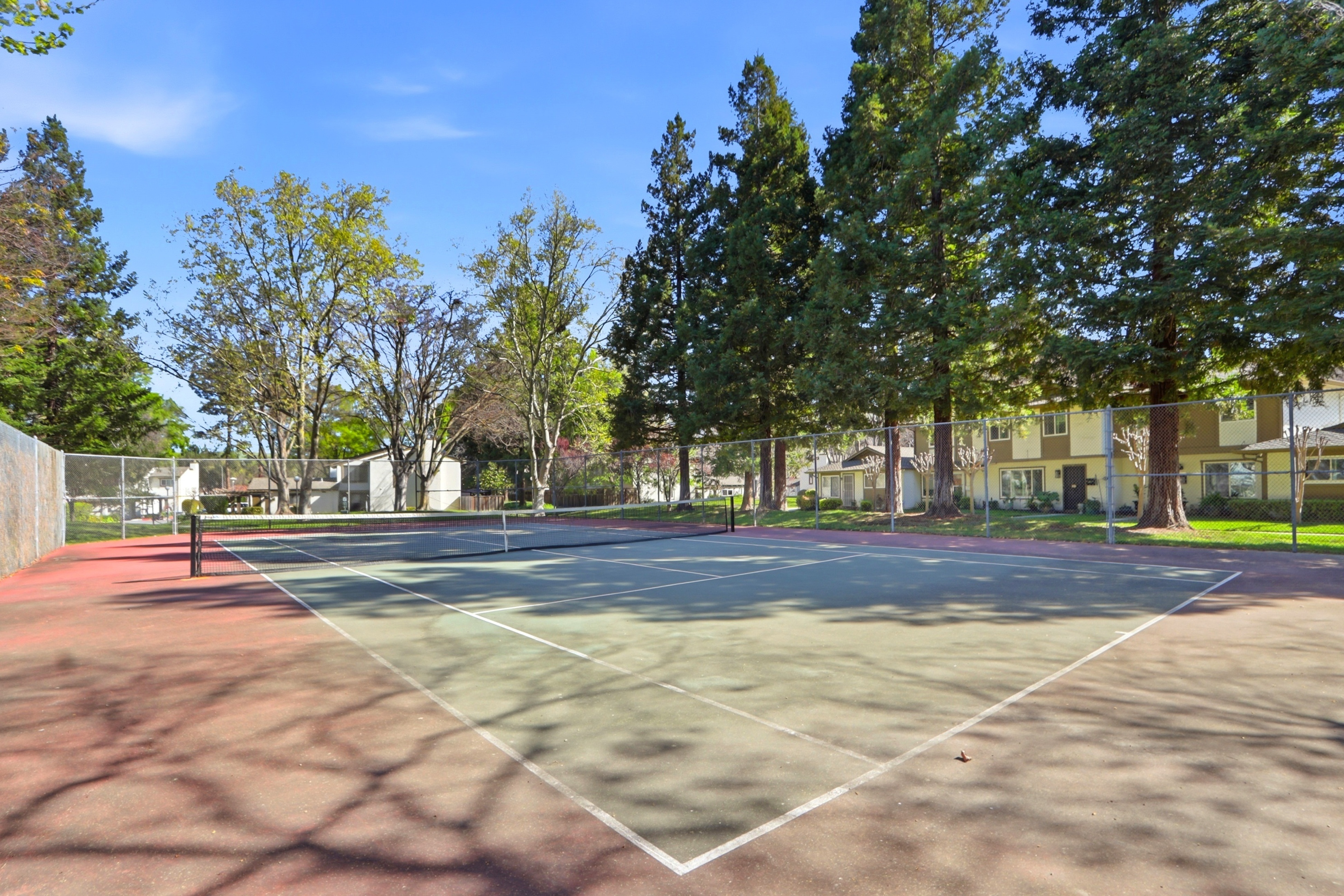 This image features a well-maintained outdoor tennis court enclosed by a chain-link fence, set within a lush, tree-lined community area. The perspective is from the baseline, looking across the court toward the net and the surrounding residential buildings in the background. The scene conveys a peaceful, active lifestyle environment with dappled sunlight filtering through the mature trees.