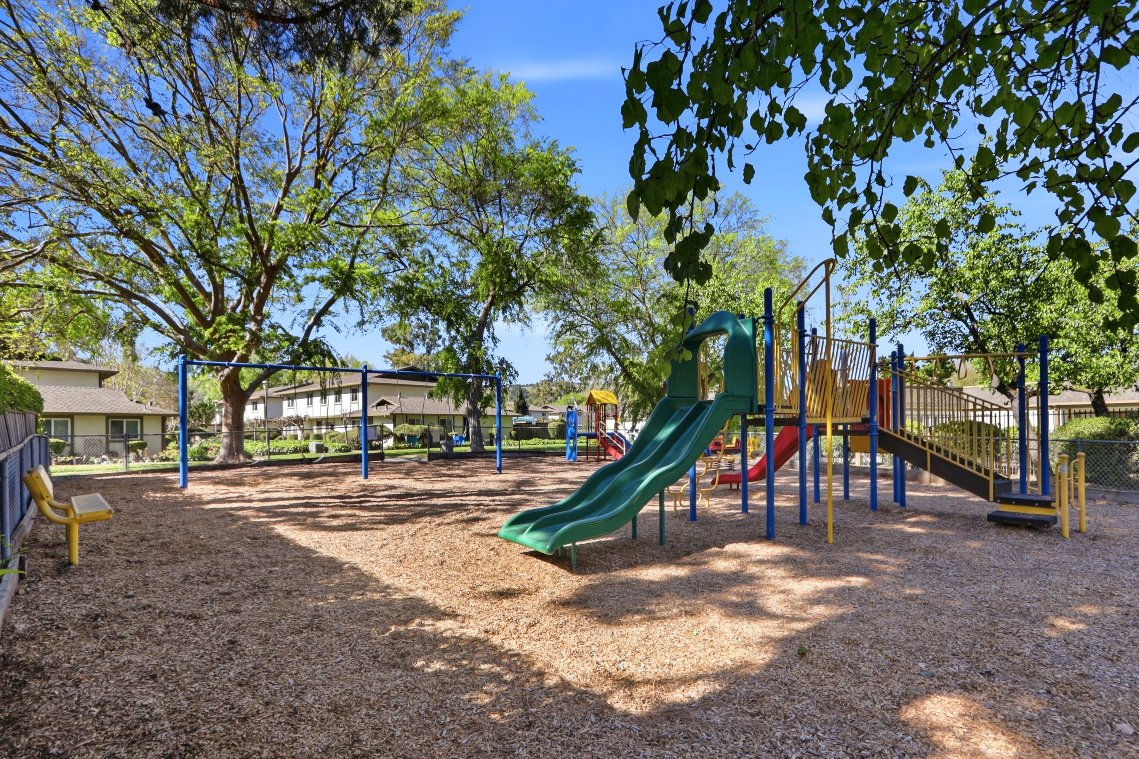 This image showcases a well-maintained community playground featuring a multi-level play structure with slides, climbing elements, and a swing set. The area is covered in wood chips and surrounded by mature trees, providing a pleasant and shaded environment for recreation. The perspective is a wide, eye-level shot that captures the entire play area, emphasizing the family-friendly amenities available to residents.