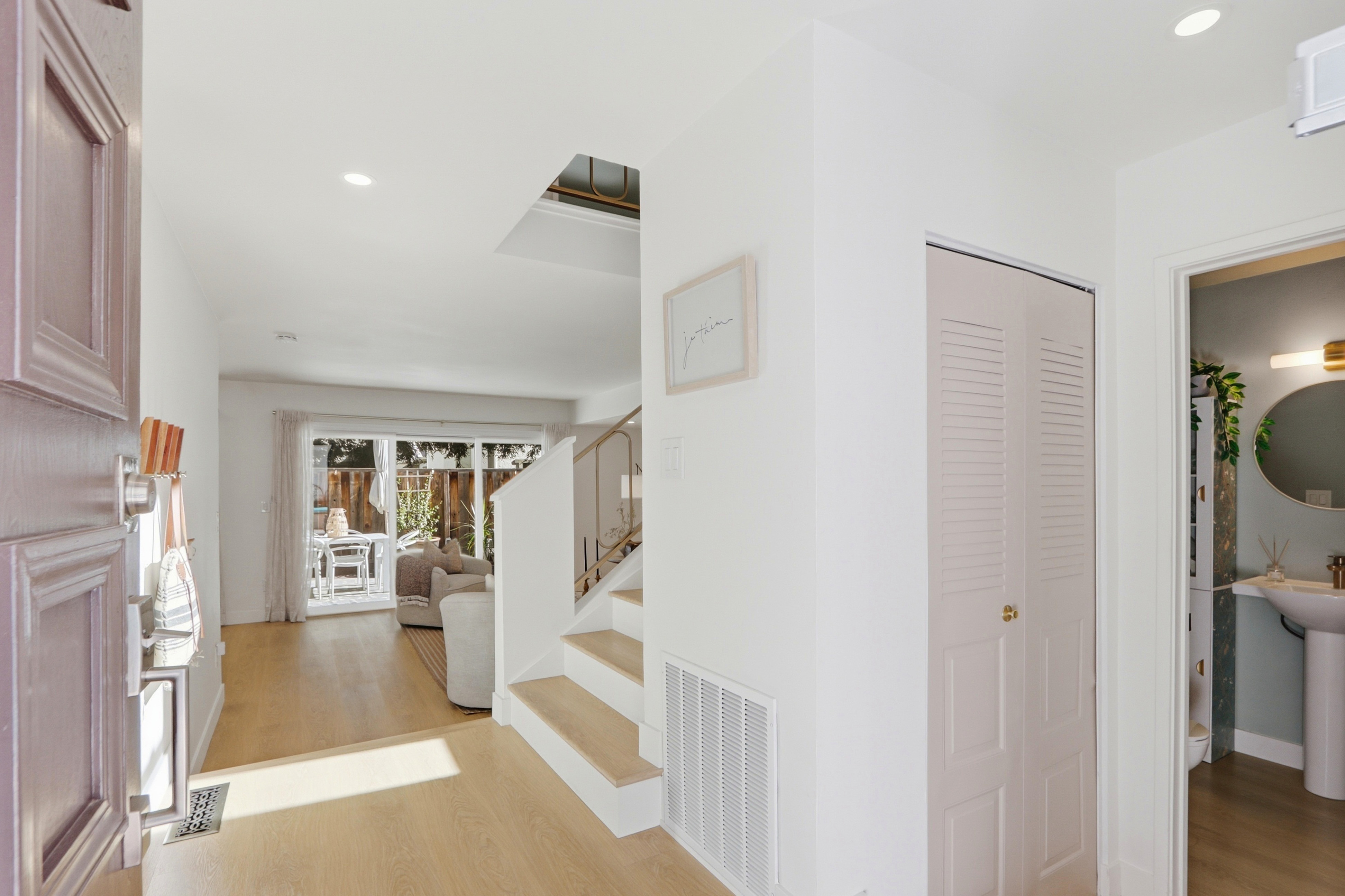 This bright and inviting entryway features light wood flooring and a clean, minimalist aesthetic. The space includes a staircase with a modern railing, a closet with louvered doors, and a glimpse into a powder room and a sunlit living area beyond. The perspective is from the front doorway, looking inward to showcase the home's open and airy floor plan.