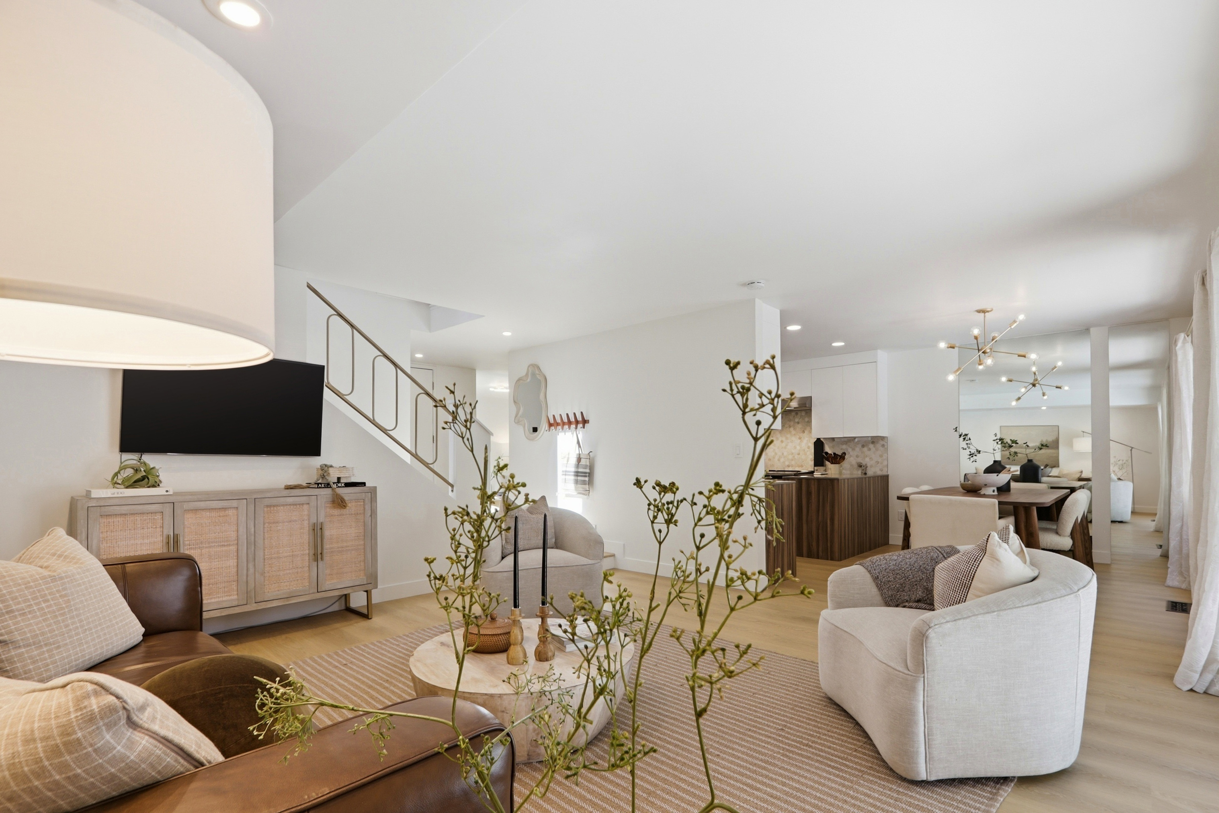 This bright and airy open-concept living room features a sophisticated blend of modern and organic textures, including a leather sofa, light-toned wood flooring, and a textured area rug. The space is anchored by a stylish media console beneath a wall-mounted television, with a staircase featuring a unique gold railing adding architectural interest. A neutral color palette and soft, layered lighting create a welcoming and cohesive atmosphere that flows seamlessly into the adjacent dining and kitchen areas.