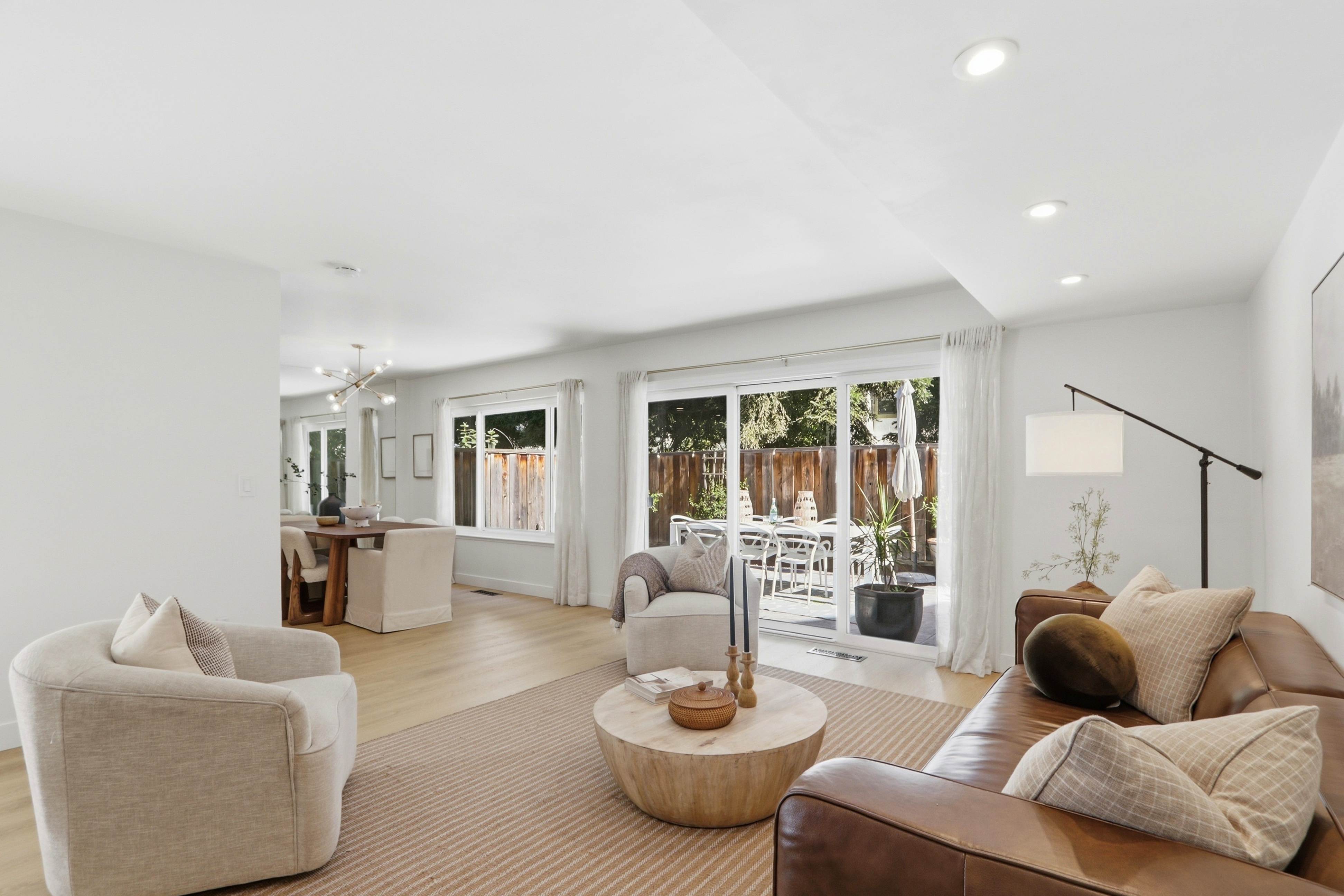 This bright and airy open-concept living room features a warm, neutral color palette with a comfortable leather sofa and plush accent chairs arranged around a rustic wooden coffee table. The space flows seamlessly into a dining area and opens up to a patio through large sliding glass doors, creating a perfect indoor-outdoor living experience. The minimalist decor and light wood flooring contribute to a clean, inviting, and modern aesthetic.