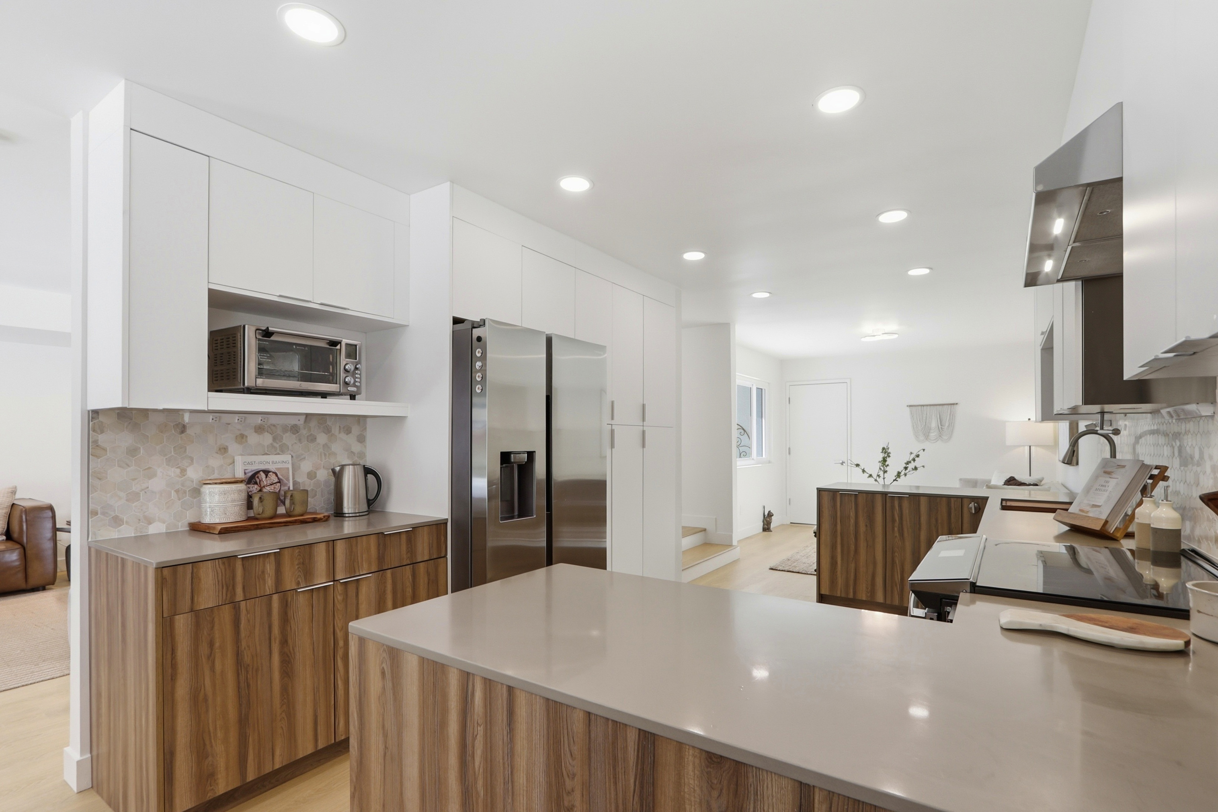This modern kitchen features a sleek, two-tone design with warm wood-grain lower cabinets and crisp white upper cabinetry. The space is equipped with stainless steel appliances, including a large refrigerator and a built-in microwave, complemented by light-colored quartz countertops and a decorative hexagonal tile backsplash. The open-concept layout flows seamlessly into the adjacent living area, creating a bright, inviting atmosphere perfect for contemporary living.