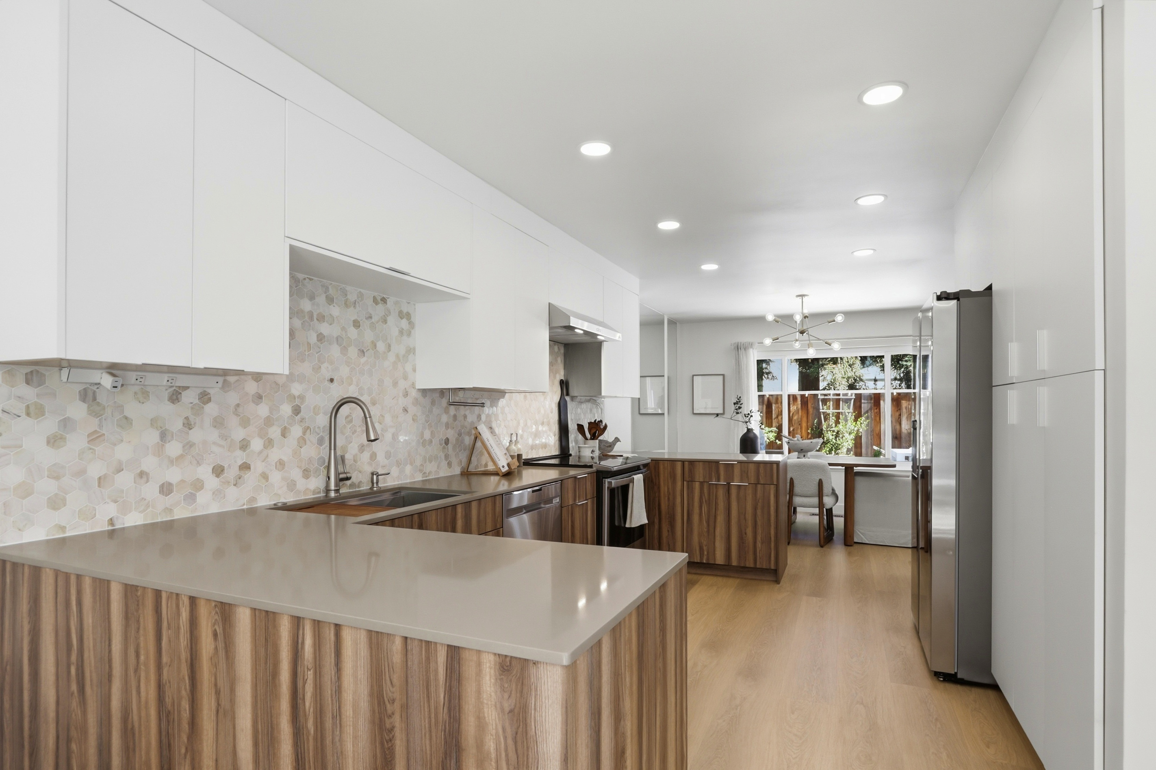 This modern, open-concept kitchen features a striking contrast between sleek white upper cabinetry and warm, wood-grain lower cabinets. The space is anchored by a smooth, light-toned quartz countertop and a unique hexagonal mosaic tile backsplash, while recessed lighting provides a bright, inviting atmosphere. The perspective looks from the kitchen workspace toward an adjacent dining area, highlighting the seamless flow and contemporary design of the home.