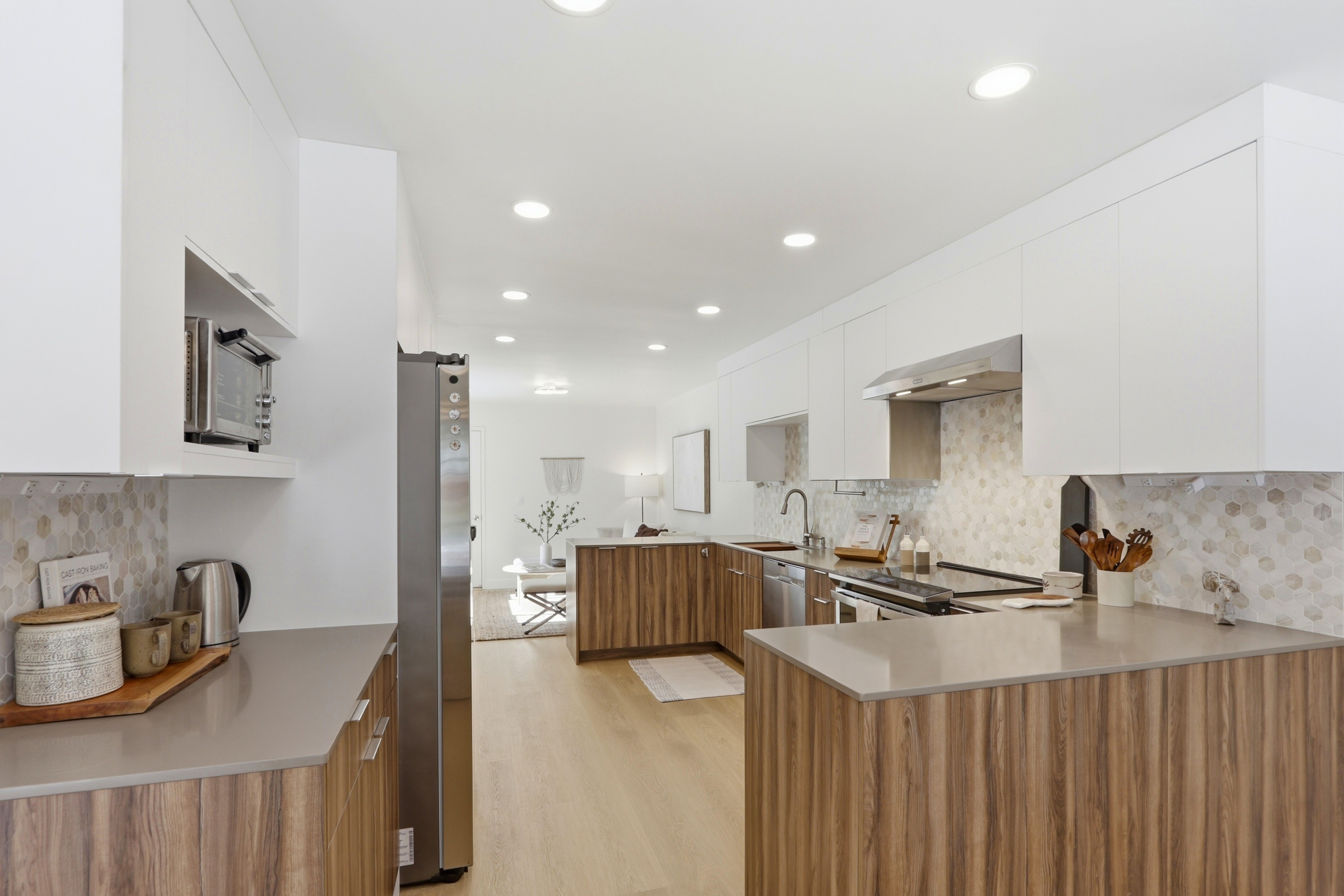 This modern, open-concept kitchen features a sleek blend of white upper cabinetry and warm wood-grain lower cabinets, complemented by neutral quartz countertops and a hexagonal tile backsplash. The space is illuminated by recessed lighting and flows seamlessly into the adjacent living area, creating an airy and inviting atmosphere. The perspective is a wide-angle shot from the kitchen entrance, showcasing the functional layout and contemporary design aesthetic.