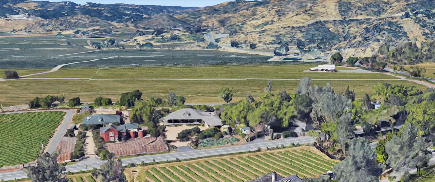 This is an aerial satellite view of a vineyard estate nestled in a scenic valley, showcasing sprawling cultivated grapevines and rolling hills in the background. The property features a primary residence, auxiliary barns, and mature landscaping, presenting a quintessential countryside lifestyle. It offers a clear perspective of the site layout, highlighting the expansive grounds and rural surroundings.