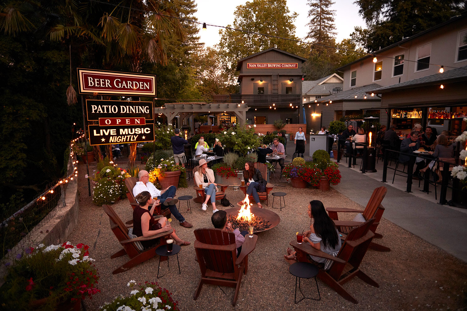 This inviting outdoor beer garden features informal lounge seating centered around a large circular fire pit, creating a cozy and social atmosphere. The patio space is accented by string lighting, vibrant flower planters, and views of the Napa Valley Brewing Company building, emphasizing its appeal as a popular community gathering spot.