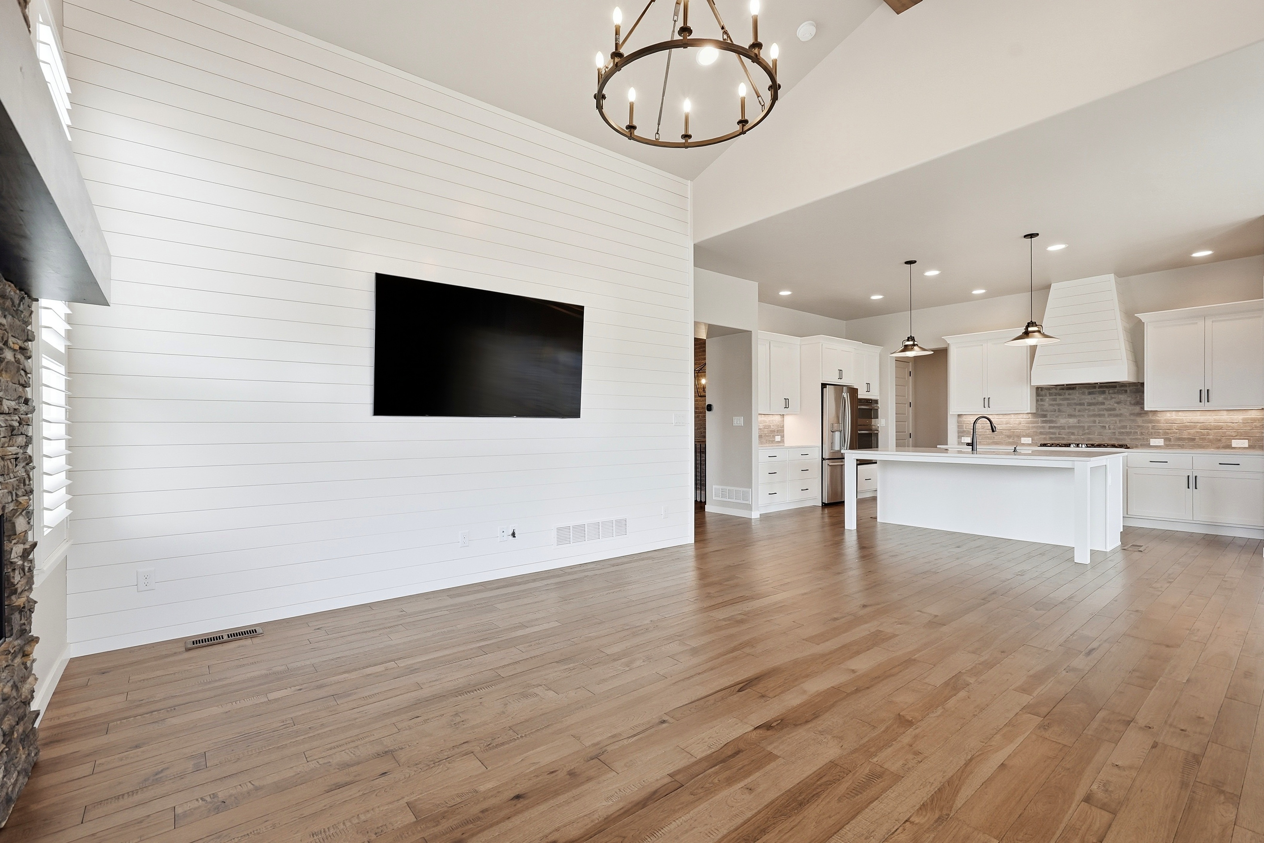 This spacious, open-concept living area features a clean, bright aesthetic highlighted by a dramatic shiplap accent wall with a mounted television and a high, vaulted ceiling anchored by a large chandelier. The hardwood floors flow seamlessly into a modern adjacent kitchen, creating a cohesive and airy living space perfect for entertaining. The combination of farmhouse-inspired textures and contemporary clean lines gives the room a sophisticated, move-in-ready feel.