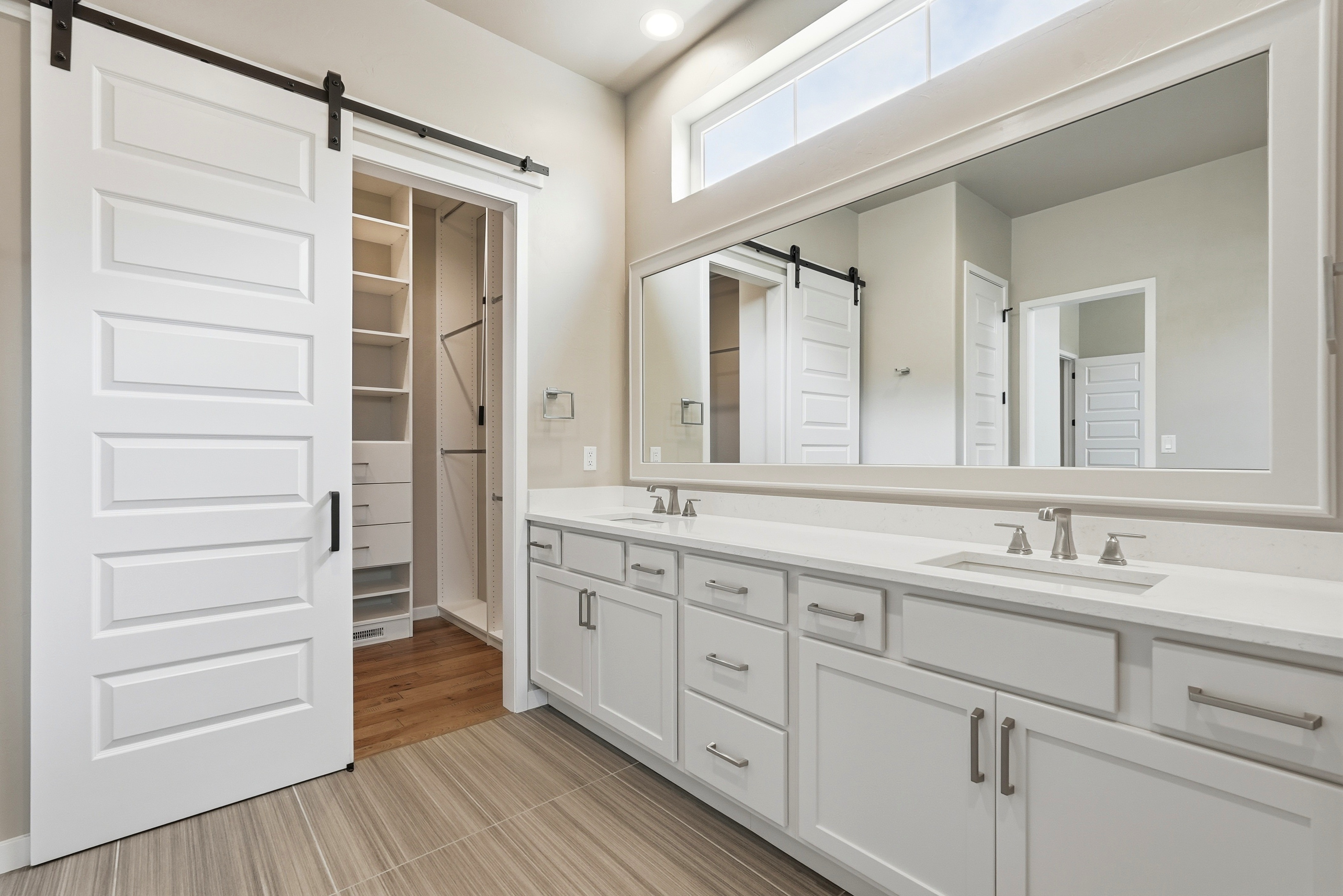 This spacious primary bathroom features a double-sink vanity with white shaker cabinetry, quartz countertops, and modern brushed nickel hardware. A large wall-to-wall mirror reflects the adjacent walk-in closet, which is accessible through a stylish sliding barn door. The room is accented by a clerestory window and sophisticated large-format tiled flooring, creating a clean, bright, and contemporary aesthetic.