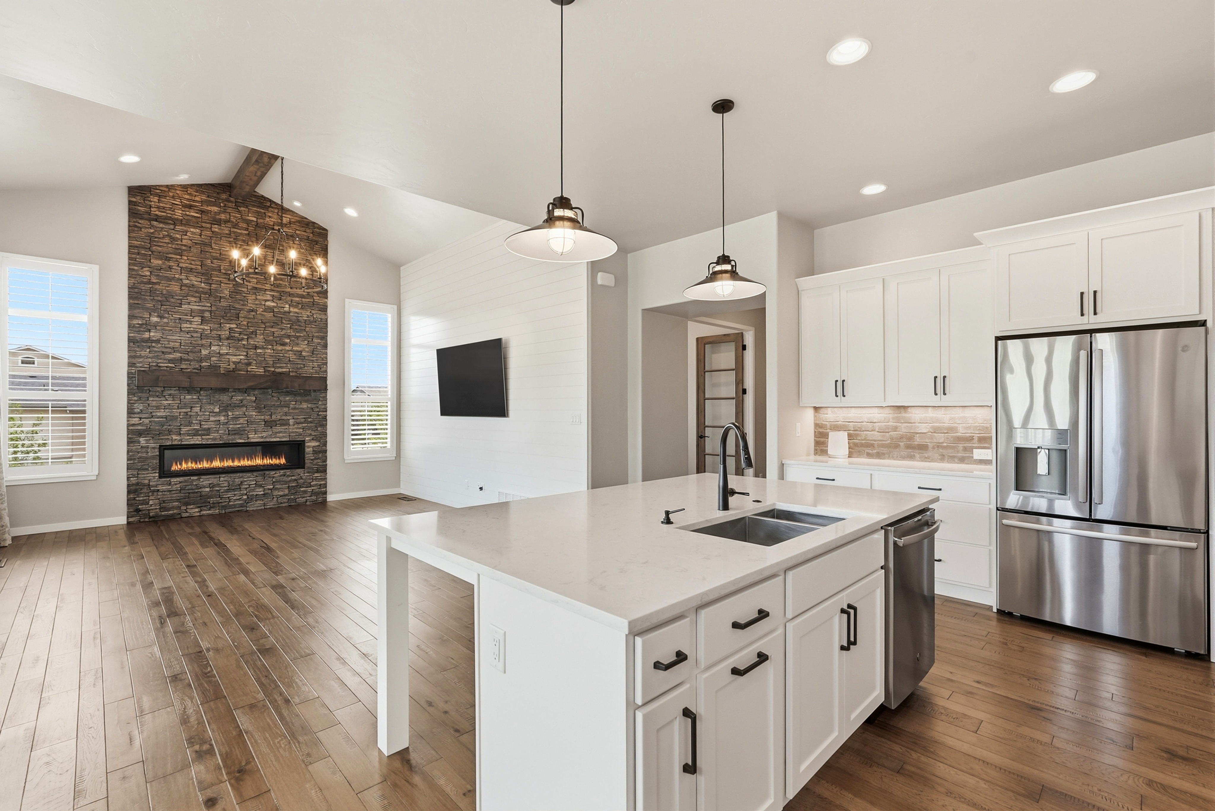 This bright, open-concept kitchen features elegant white cabinetry, a spacious quartz-topped island with a farmhouse-style faucet, and high-end stainless steel appliances. Adjacent to the kitchen is a welcoming living area anchored by a striking stone fireplace, creating a seamless and sophisticated flow between spaces. The combination of warm wood flooring, crisp white walls, and modern pendant lighting gives the home a refined, transitional aesthetic.
