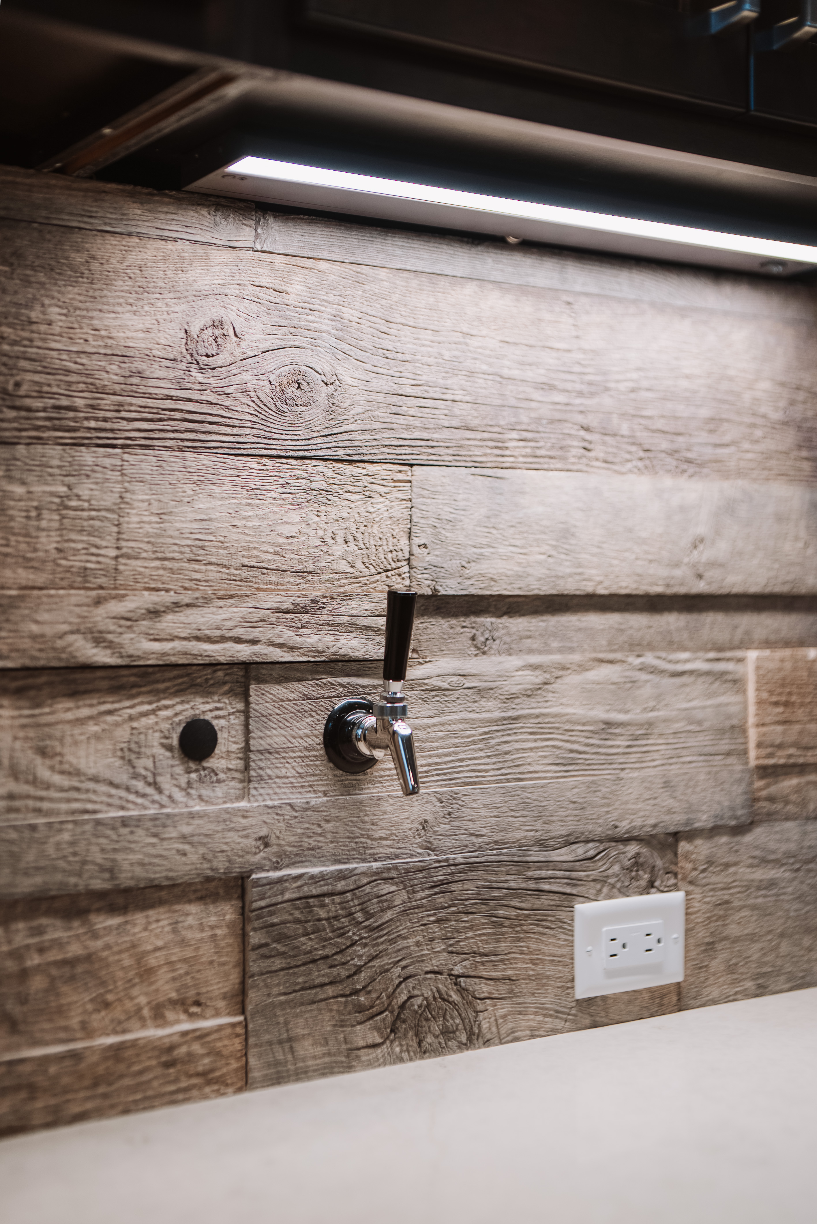 This detail shot highlights a modern beer tap installation mounted directly onto a rustic, reclaimed wood-plank backsplash. The area is accented by sleek under-cabinet lighting and a standard electrical outlet, showcasing a unique and well-executed custom home feature. The textured wood paired with the polished chrome finish of the tap creates a stylish, high-end recreational aesthetic.
