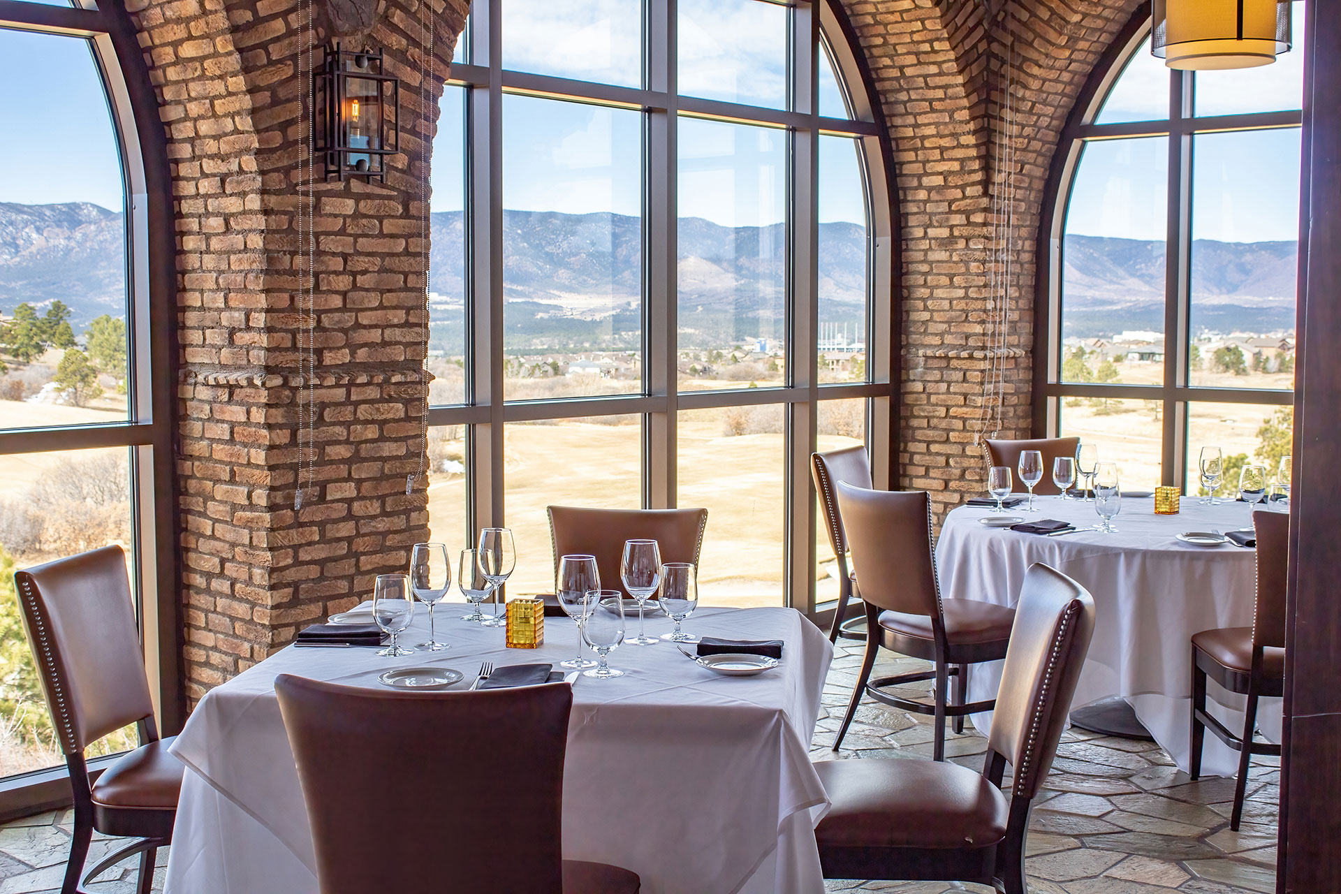 This elegant dining room features rustic brick arches and floor-to-ceiling windows that provide panoramic views of the surrounding mountainous landscape. The space is furnished with set tables featuring crisp white linens, high-backed leather chairs, and sophisticated glassware. Its combination of natural architectural textures and expansive scenic vistas creates a refined and inviting atmosphere for upscale dining.