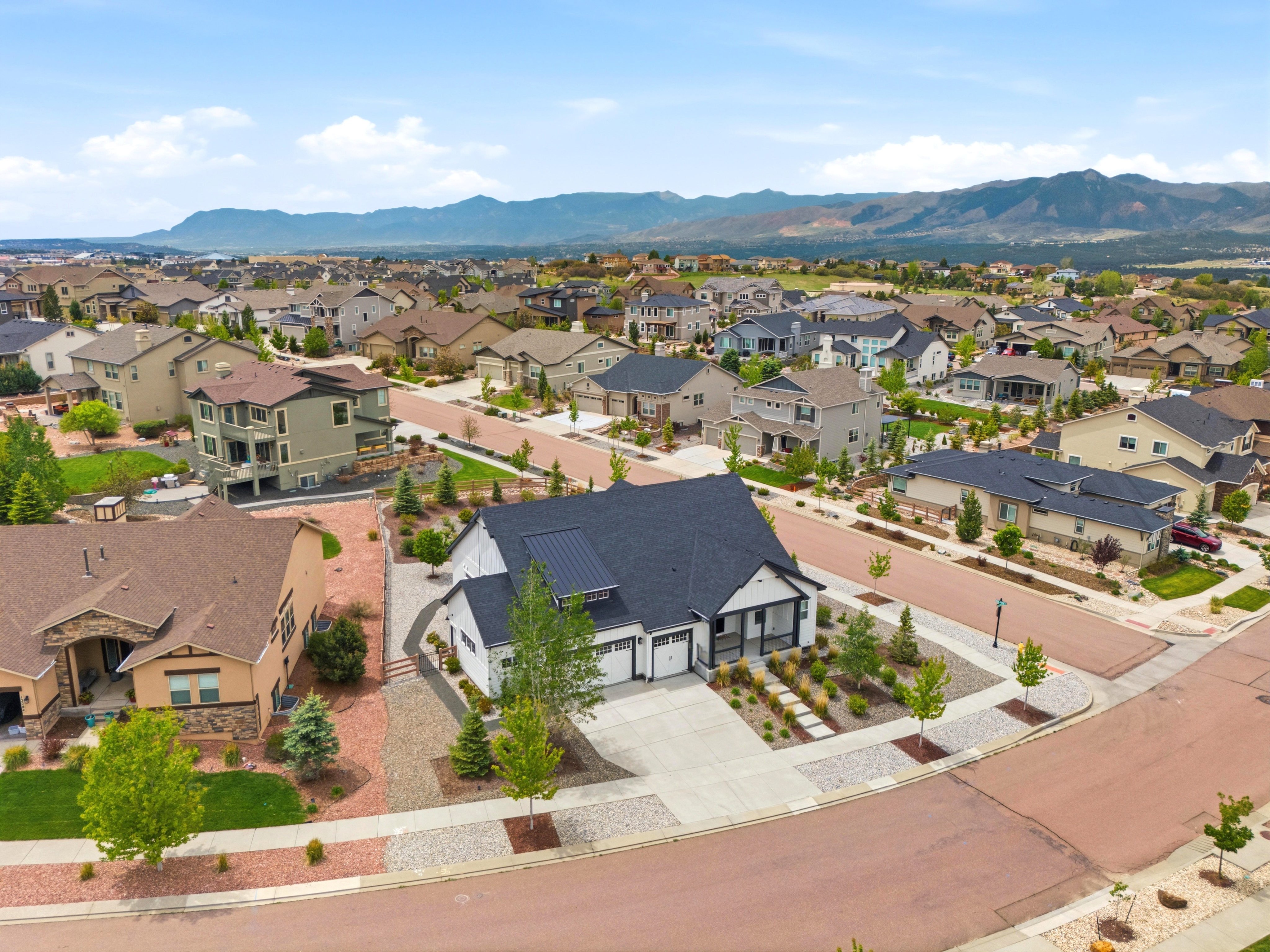 This aerial view captures a sunny suburban neighborhood nestled at the foot of a mountain range. The image highlights a modern, white farmhouse-style home with a dark roof and a spacious concrete driveway as its focal point, surrounded by well-maintained landscaping and neighbouring houses. The overall impression is one of a clean, peaceful, and family-friendly residential community.