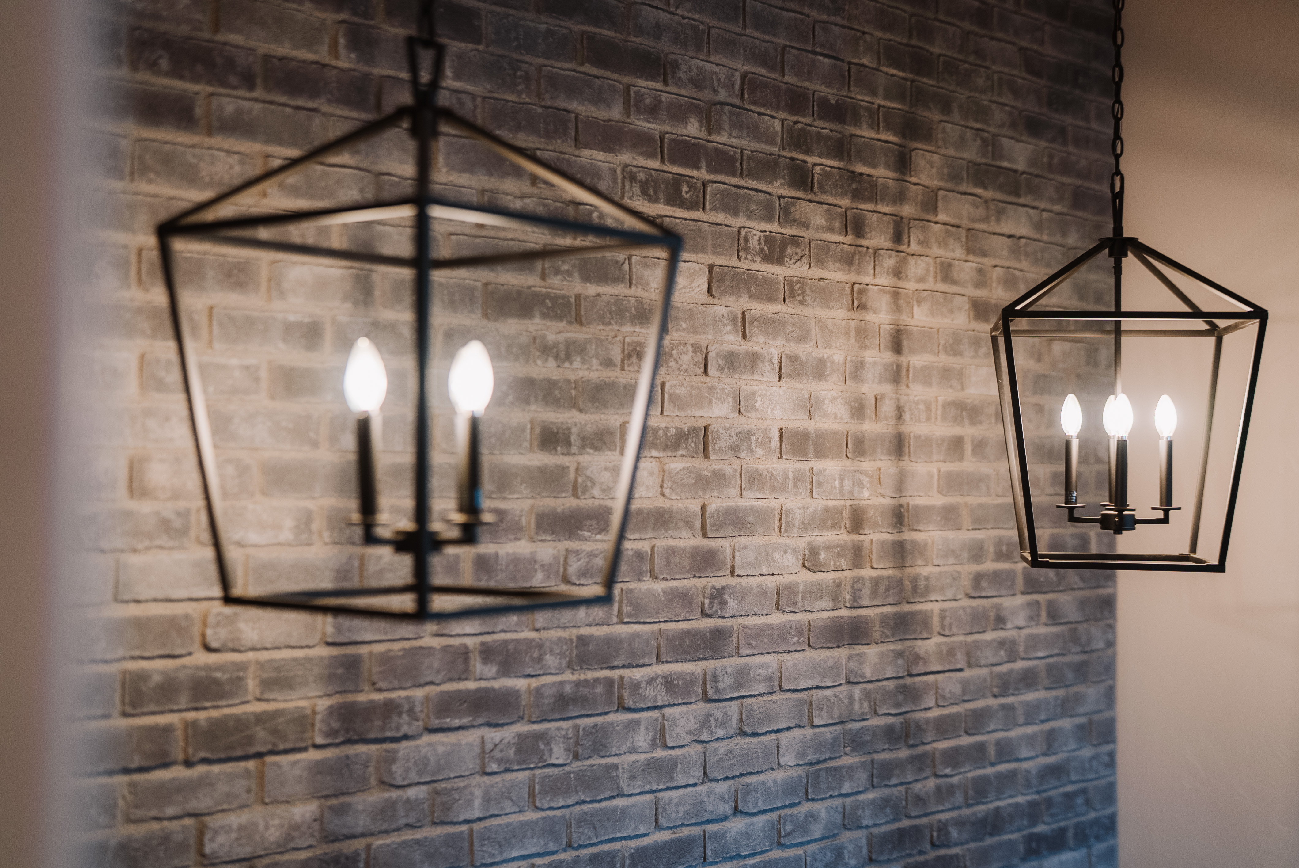 This image highlights a pair of elegant, modern geometric pendant chandeliers featuring a sleek black metal frame. These fixtures are set against a rustic, textured gray brick wall, creating a sophisticated contrast that adds visual depth and character to the interior space.