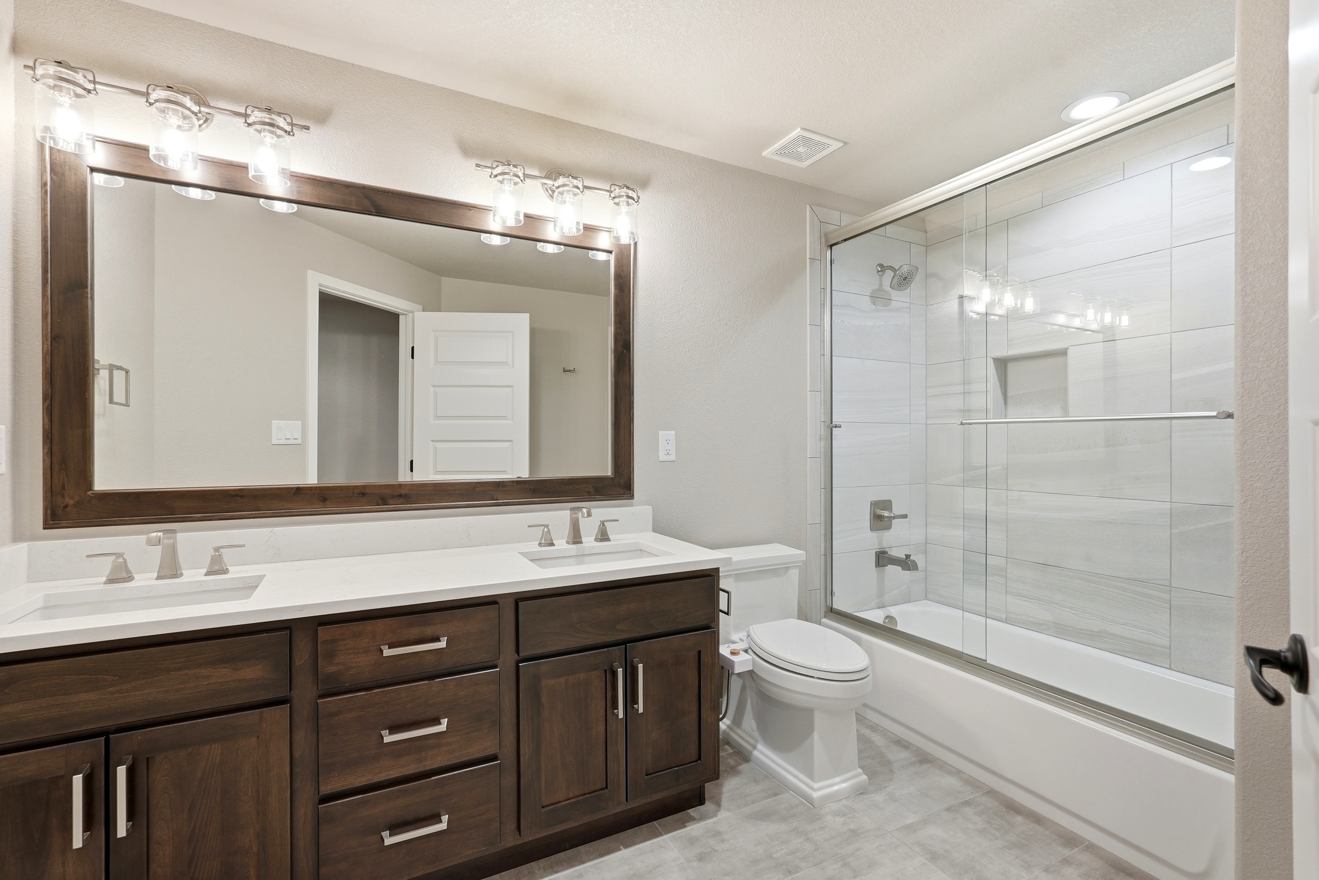 This clean and modern bathroom features a double-sink vanity with dark wood cabinetry, white quartz countertops, and a large framed mirror with stylish light fixtures. Adjacent to the vanity is a toilet and a tiled, glass-enclosed bathtub-shower combination. The neutral color palette and functional layout create a welcoming and contemporary space suitable for guests.