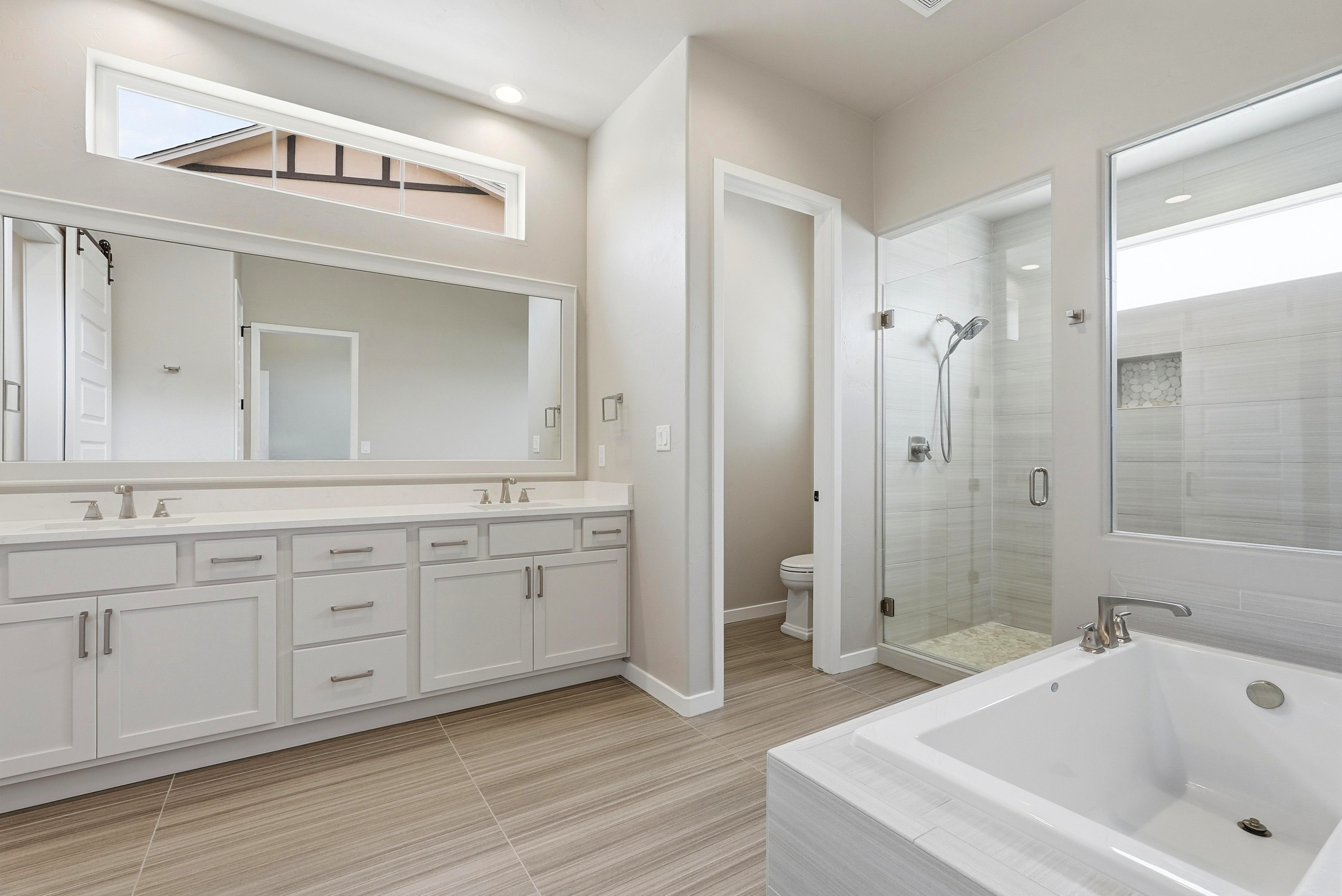 This modern primary bathroom features a double-sink vanity with clean white cabinetry and quartz countertops, complemented by contemporary brushed-nickel fixtures. A separate glass-enclosed shower and a soaking tub create a spa-like atmosphere, while neutral, wood-look tile flooring and high ceilings with transom windows provide an airy and sophisticated feel.