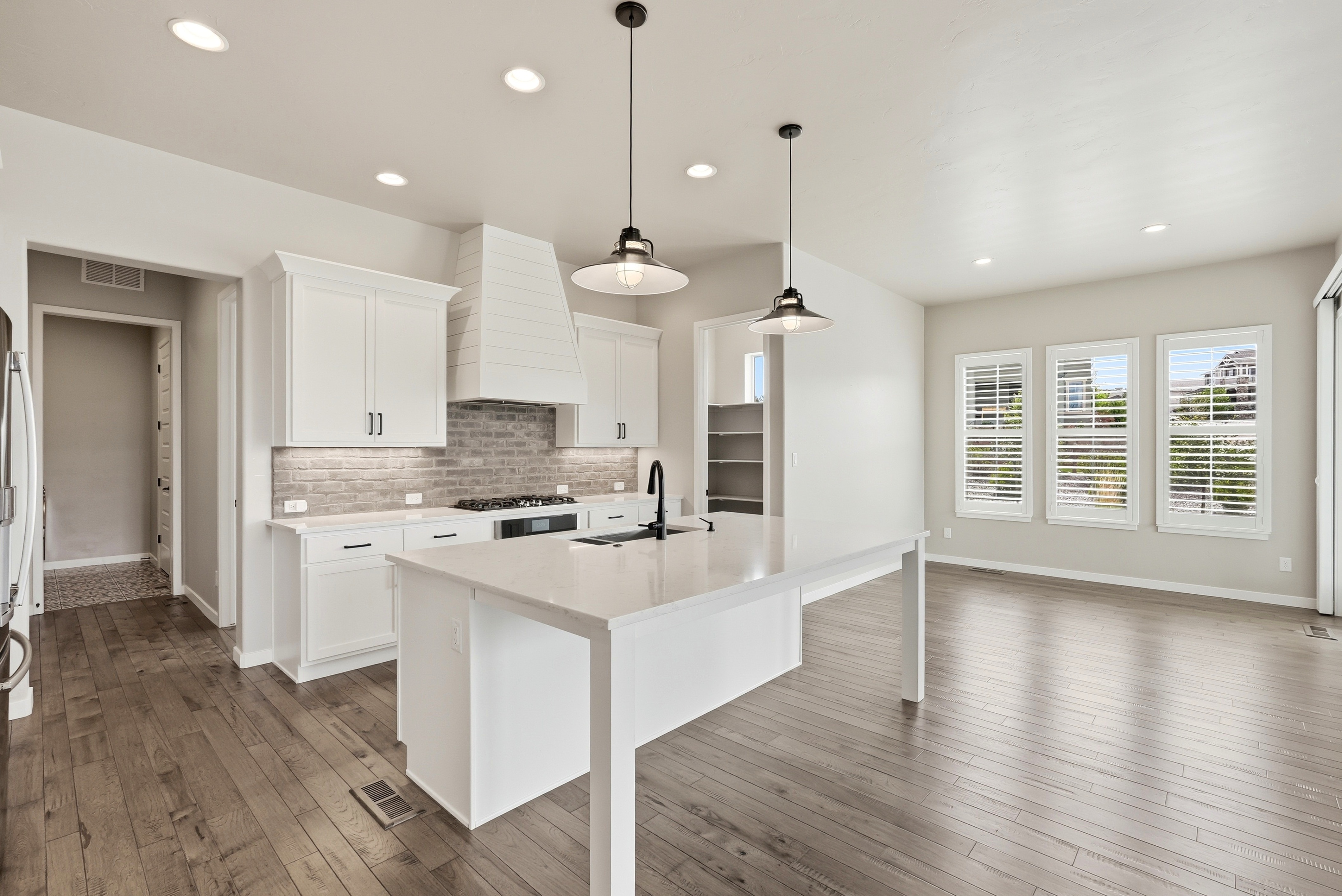This bright, contemporary kitchen showcases clean lines with crisp white cabinetry, light quartz countertops, and a stylish gray brick backsplash. The spacious center island features a deep sink and dramatic black fixtures, complemented by dual industrial-style pendant lights above. Open-concept transitions and warm hardwood flooring give the home a polished, inviting feel, perfect for modern living.