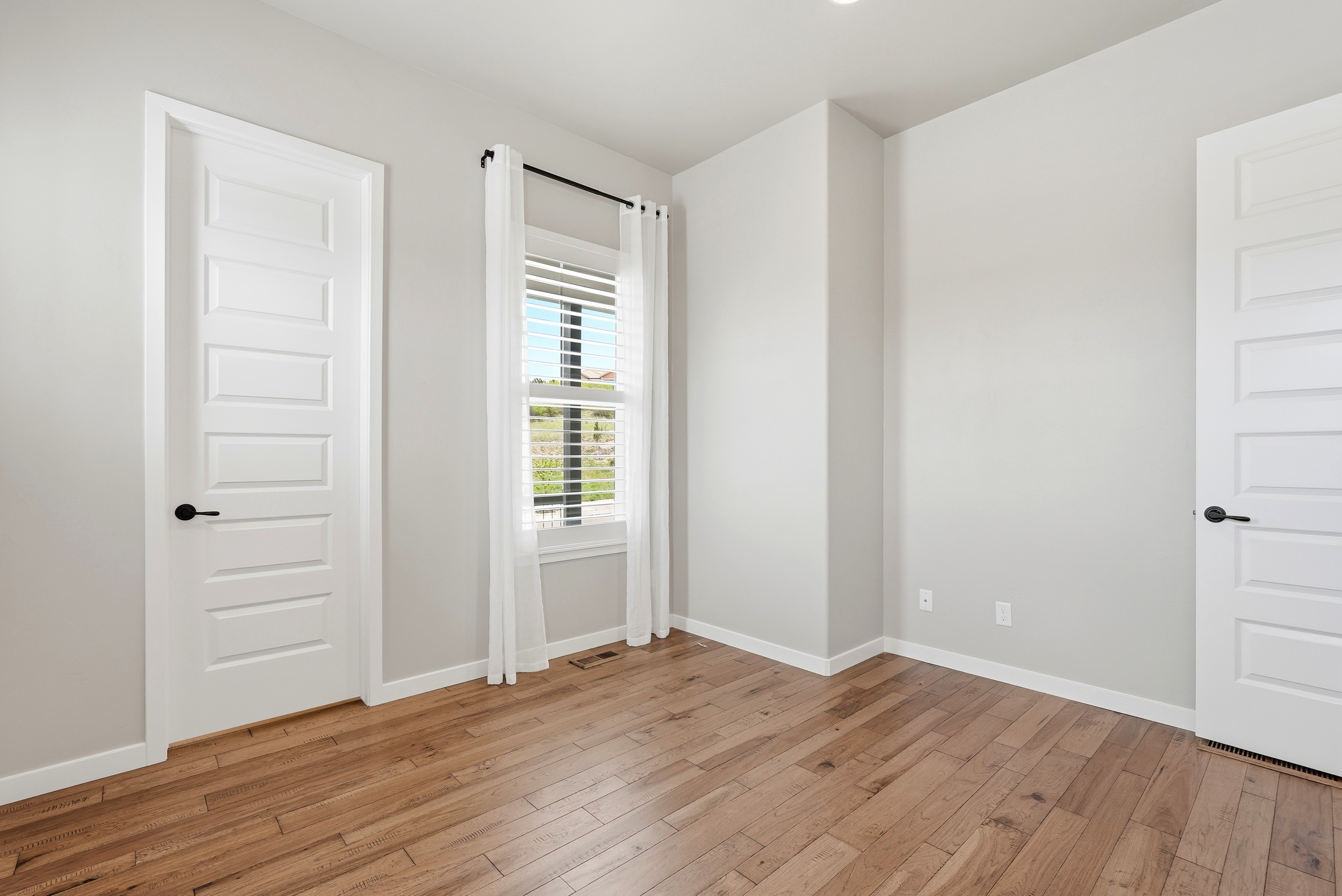 This is a clean, neutral bedroom with light-toned walls and warm-toned hardwood flooring throughout. The room features two white interior doors, a window adorned with white curtains and blinds, and minimalist architectural details. The space feels bright, inviting, and move-in ready.