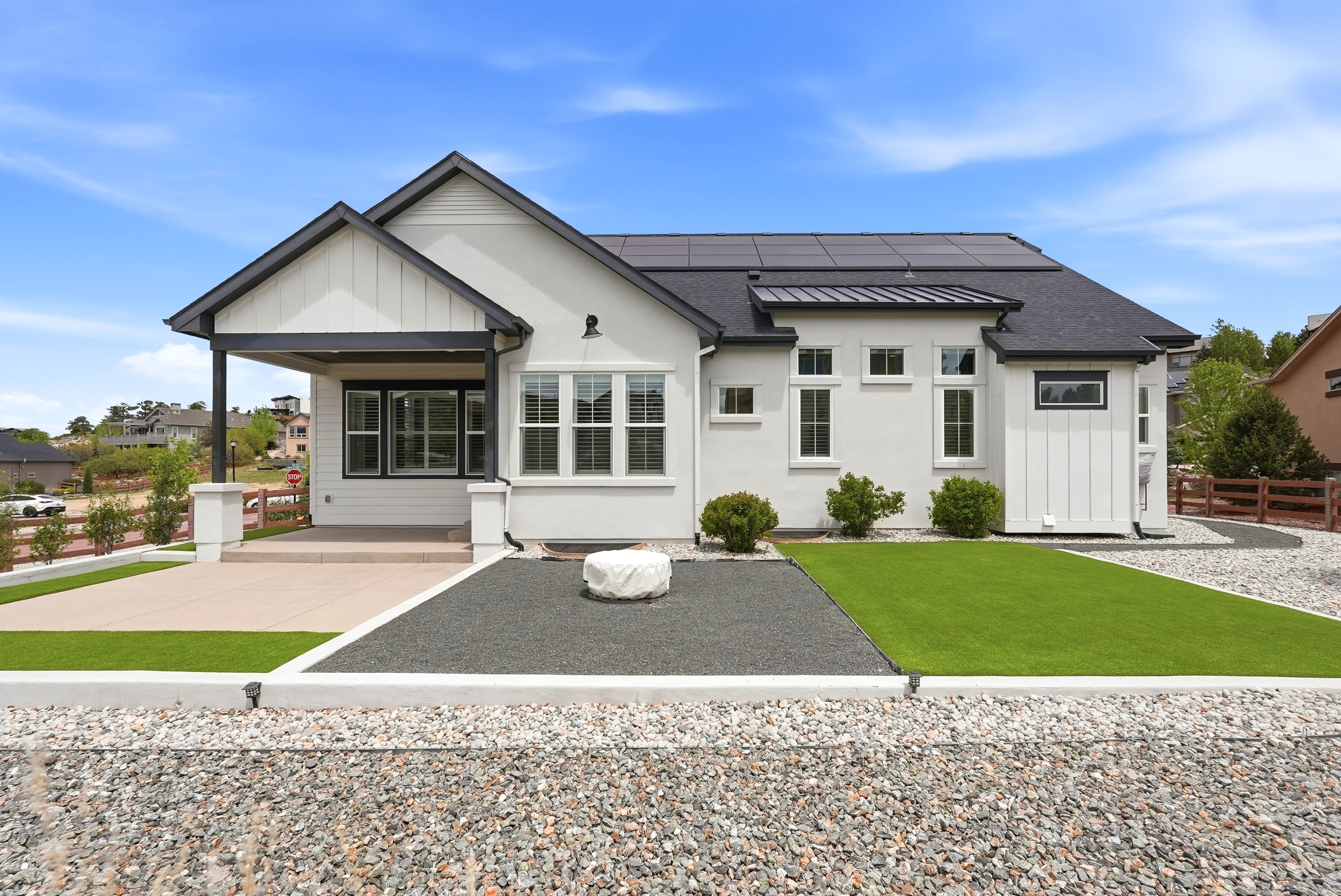 This modern single-story residence showcases a contemporary farmhouse aesthetic with clean white siding, dark window frames, and a combination of shingle and metal roofing including integrated solar panels. The backyard features a professional landscaped mix of artificial turf, a decorative gravel patio area, and a covered porch, creating a tidy and low-maintenance outdoor living space. The home sits on a spacious lot that blends seamlessly with the surrounding neighborhood.