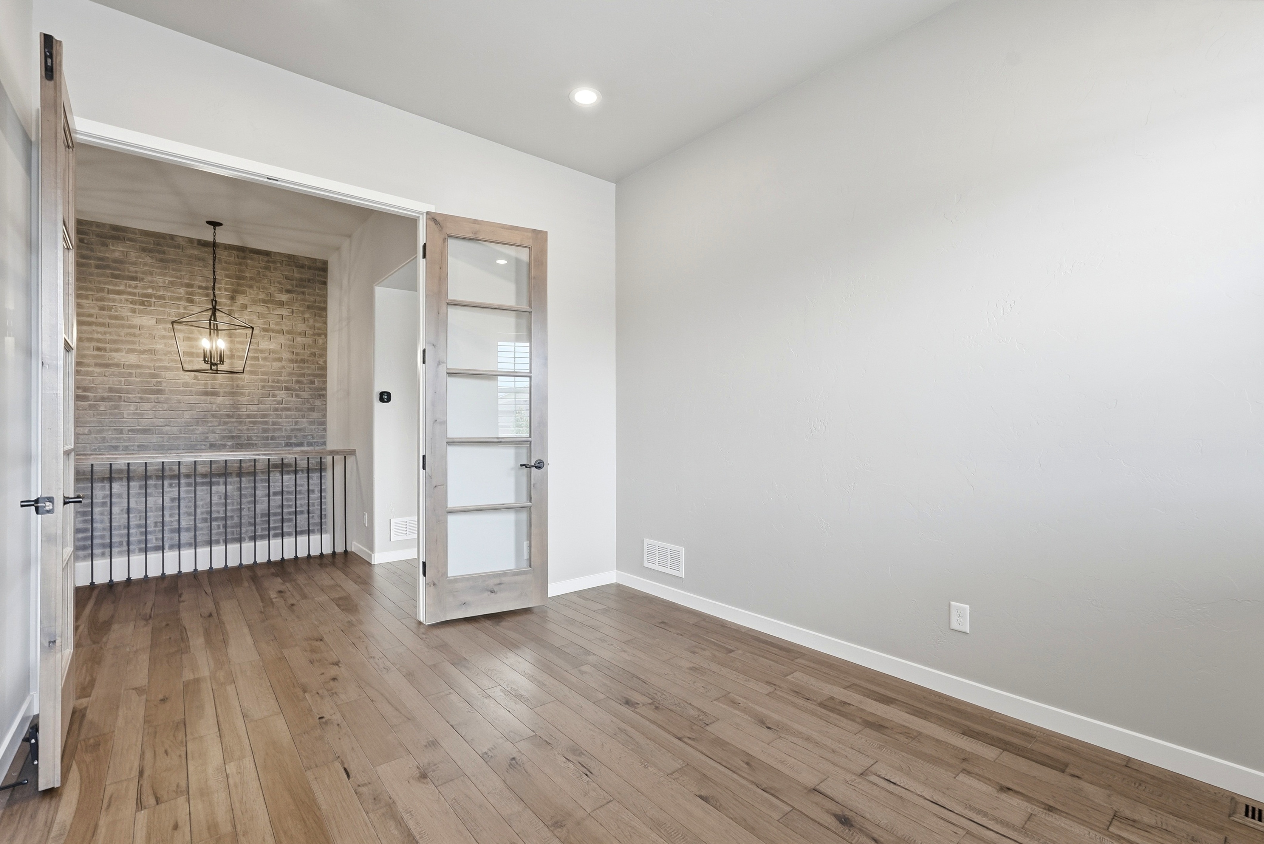 This bright, modern room features light-colored hardwood flooring and neutral walls, making it a versatile space perfect for a home office or flex room. Through the elegant French glass doors, a secondary area is visible with an exposed brick wall, a contemporary geometric pendant light, and a decorative railing, adding unique architectural character to the home.