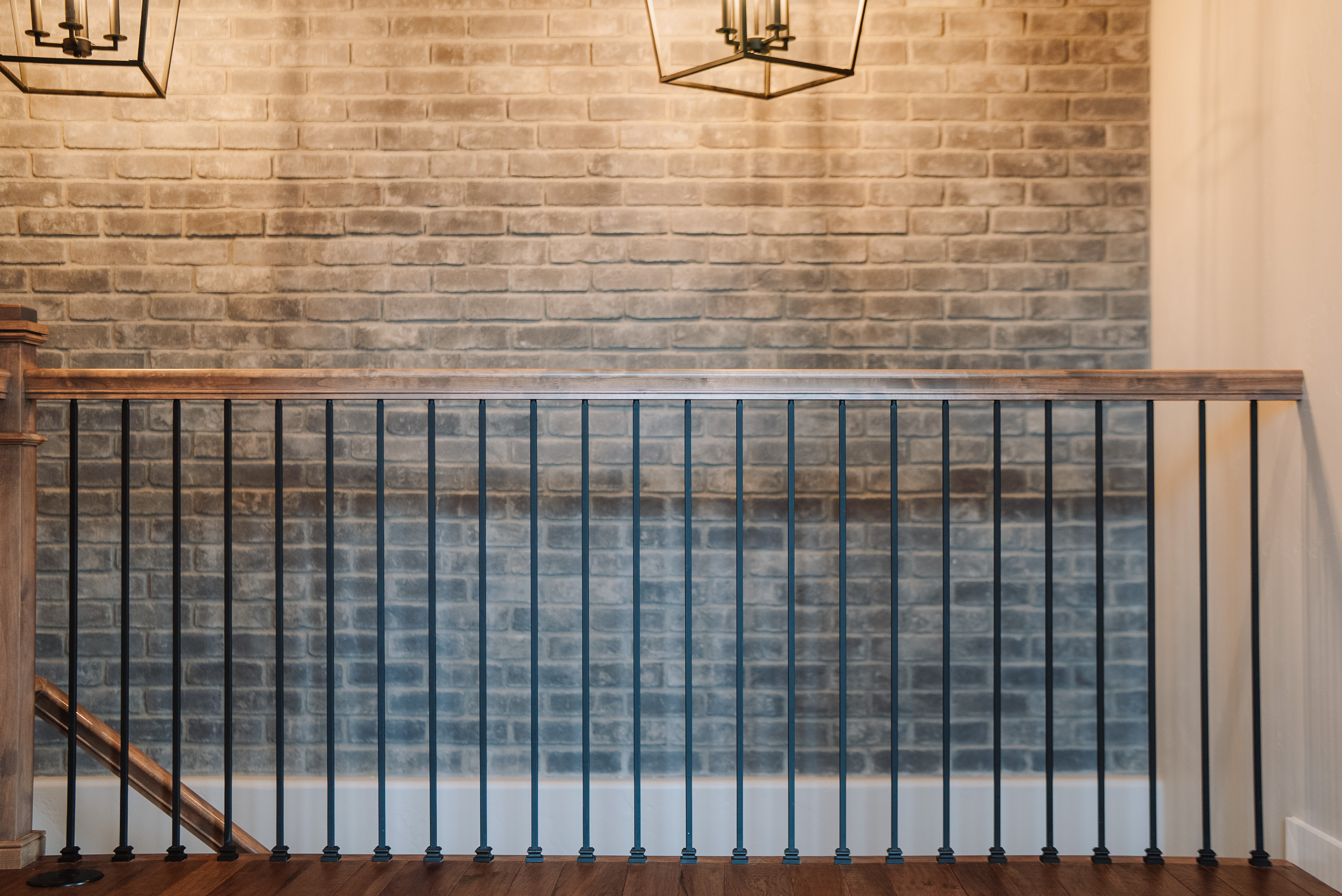 This image highlights a modern staircase railing featuring sleek black metal balusters topped by a warm-toned wooden handrail. In the background, a textured gray brick wall adds a rustic yet contemporary charm to the space. The inclusion of decorative pendant light fixtures at the top suggests a well-designed and stylish interior design aesthetic.