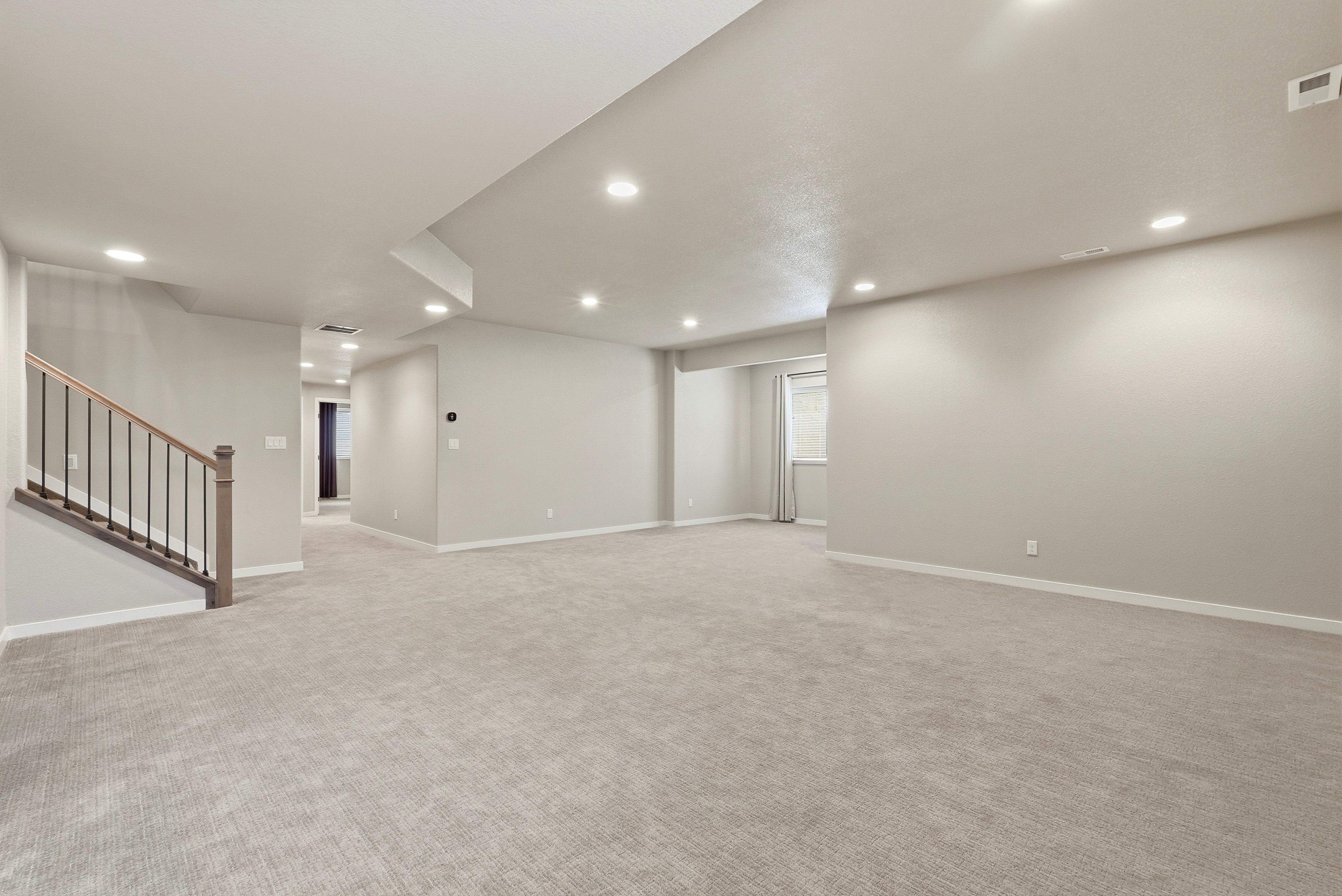 This spacious, unfinished basement offers a versatile open-concept layout with neutral-colored walls and plush carpeting, creating a bright and inviting multi-purpose space. Key architectural features include recessed lighting throughout, a clean stairwell with wood accents and metal railings, and a secondary recessed area with a window providing natural light. The neutral palette provides a perfect canvas for customization, making it an ideal area for a recreation room, home gym, or entertainment space.