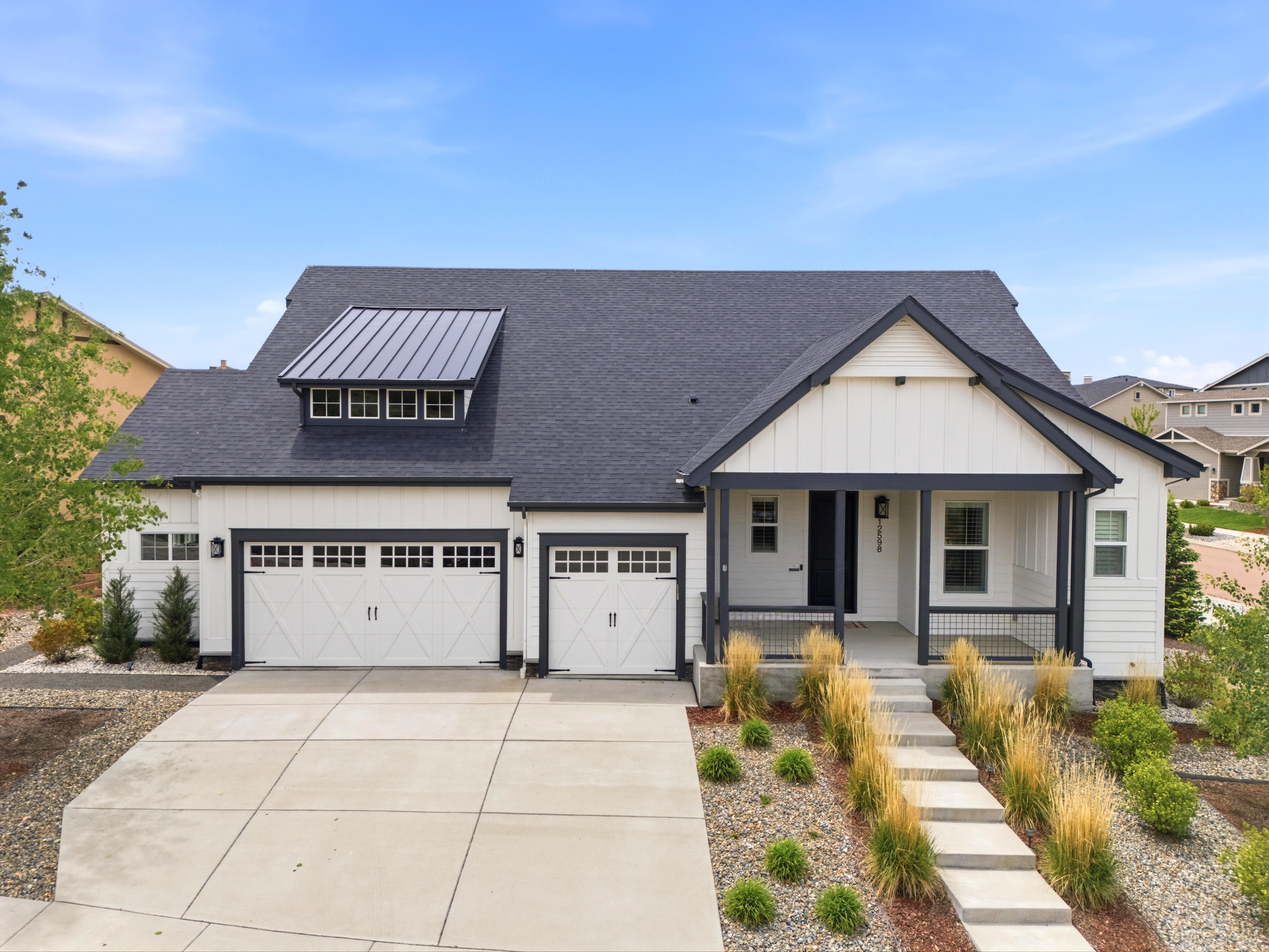 This modern farmhouse-style home features a striking combination of white board-and-batten siding and dark trim elements. The facade includes a three-car garage, a cozy covered front porch, and a metal roof accent over the garage windows, all set against a professionally landscaped lot with ornamental grasses and gravel beds. The property offers excellent curb appeal with a clean, contemporary aesthetic.