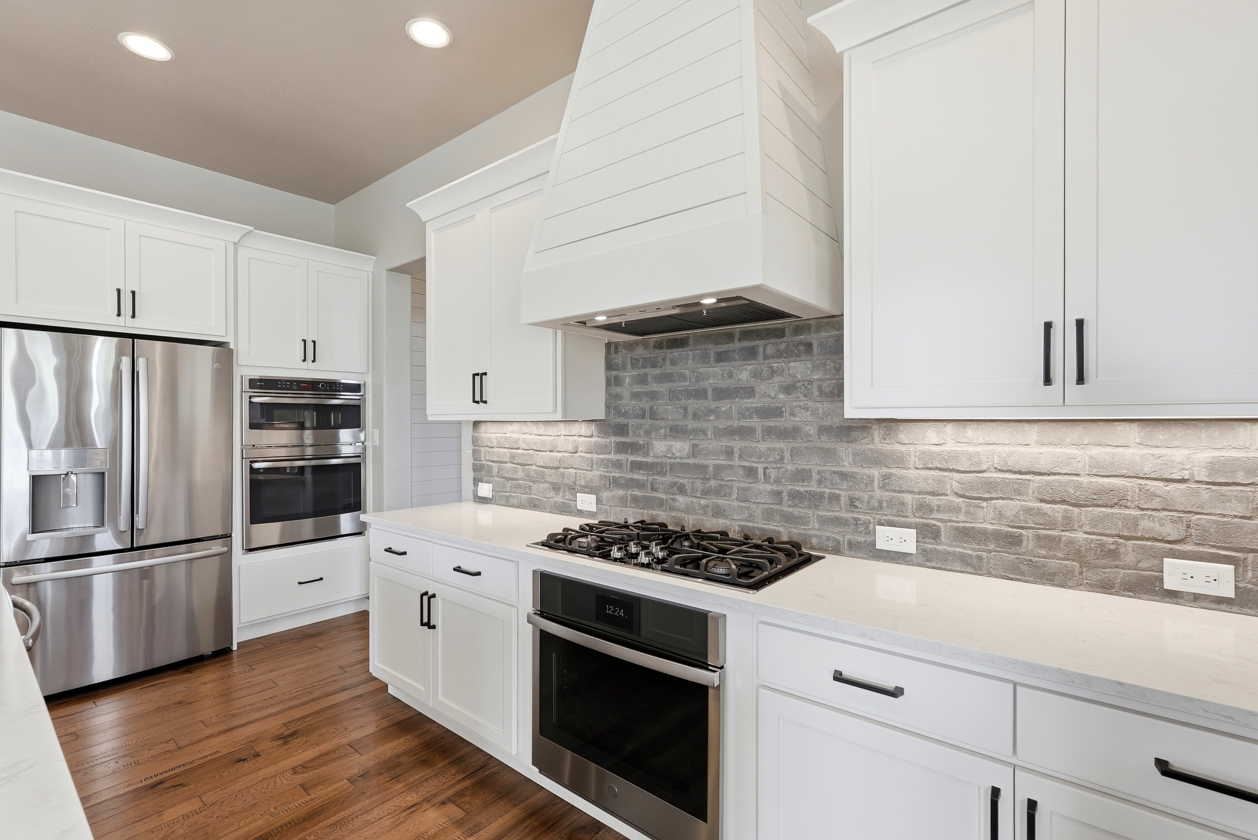This bright, contemporary kitchen features white shaker-style cabinetry contrasted by a rustic, grey brick tile backsplash and warm wood flooring. It is equipped with premium stainless steel built-in appliances, including a double oven and gas cooktop, all beneath a custom range hood. The sleek quartz countertops and modern lighting fixtures create a sophisticated yet inviting space for culinary preparation.