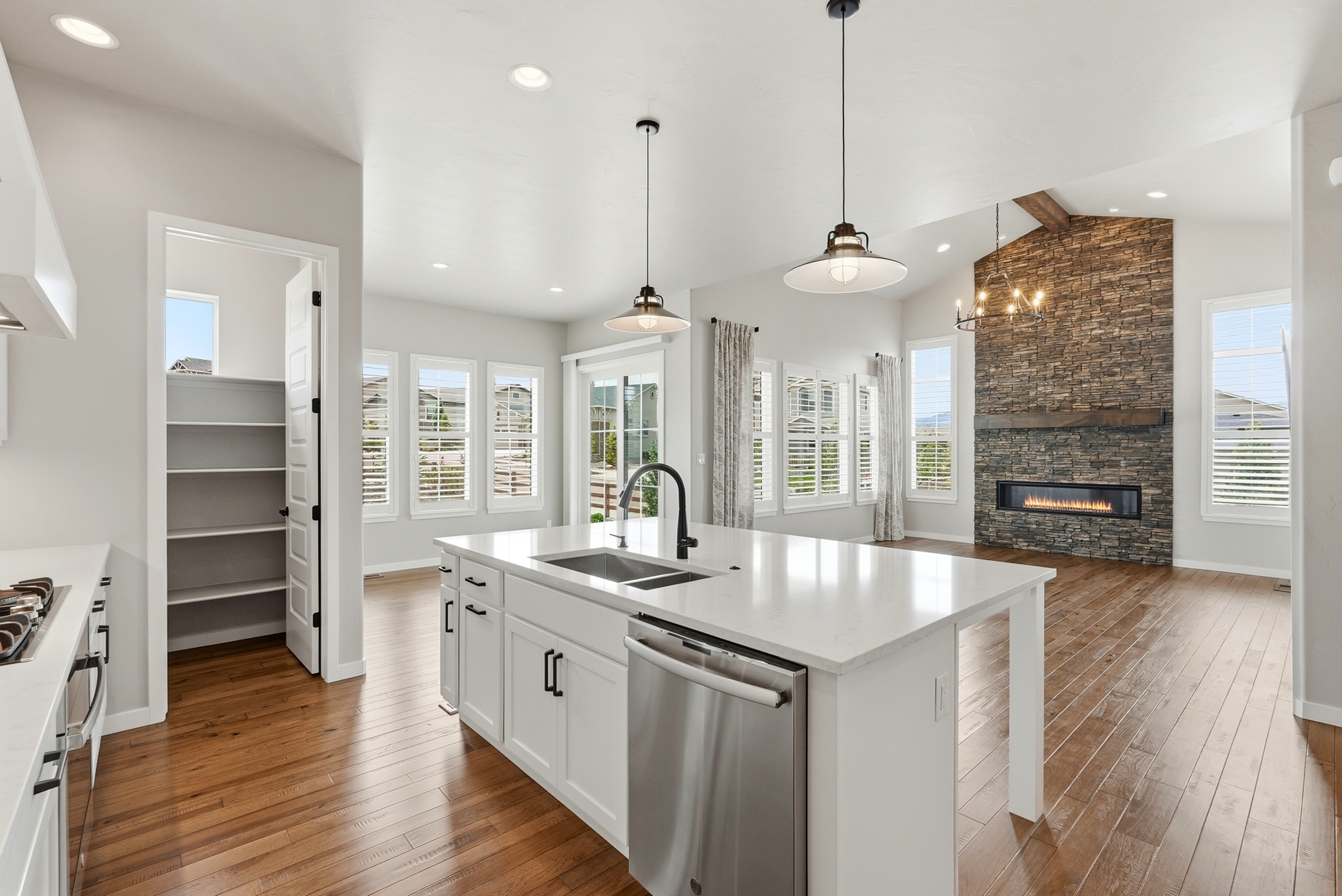 This bright and airy kitchen features white shaker-style cabinetry, quartz countertops, and a large central island with an integrated stainless steel dishwasher and sink. The open-concept design flows seamlessly into a living area highlighted by a stunning floor-to-ceiling stone fireplace and rich hardwood flooring throughout. A walk-in pantry with built-in shelving is conveniently tucked to the side, adding practical storage to the sophisticated modern aesthetic.