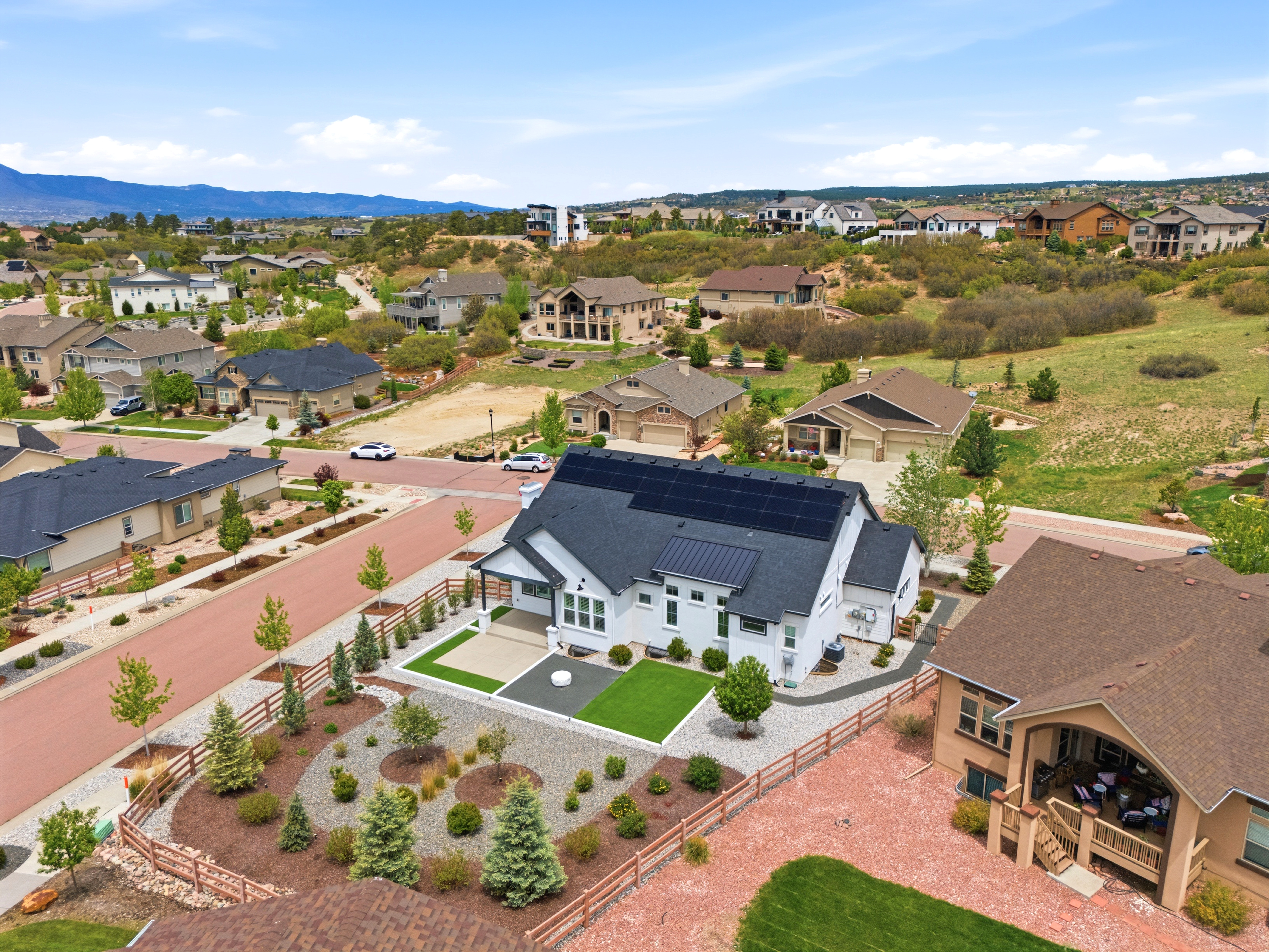 This aerial view captures a contemporary residential neighborhood featuring a centerpiece white farmhouse-style home with black roof accents and prominent solar panels. The property showcases a meticulously landscaped yard with designated low-maintenance rock gardens, artificial turf, and young evergreen plantings enclosed by clean wood fencing. The surrounding area displays a well-planned development with cohesive architectural styles and scenic mountainous vistas in the distance.