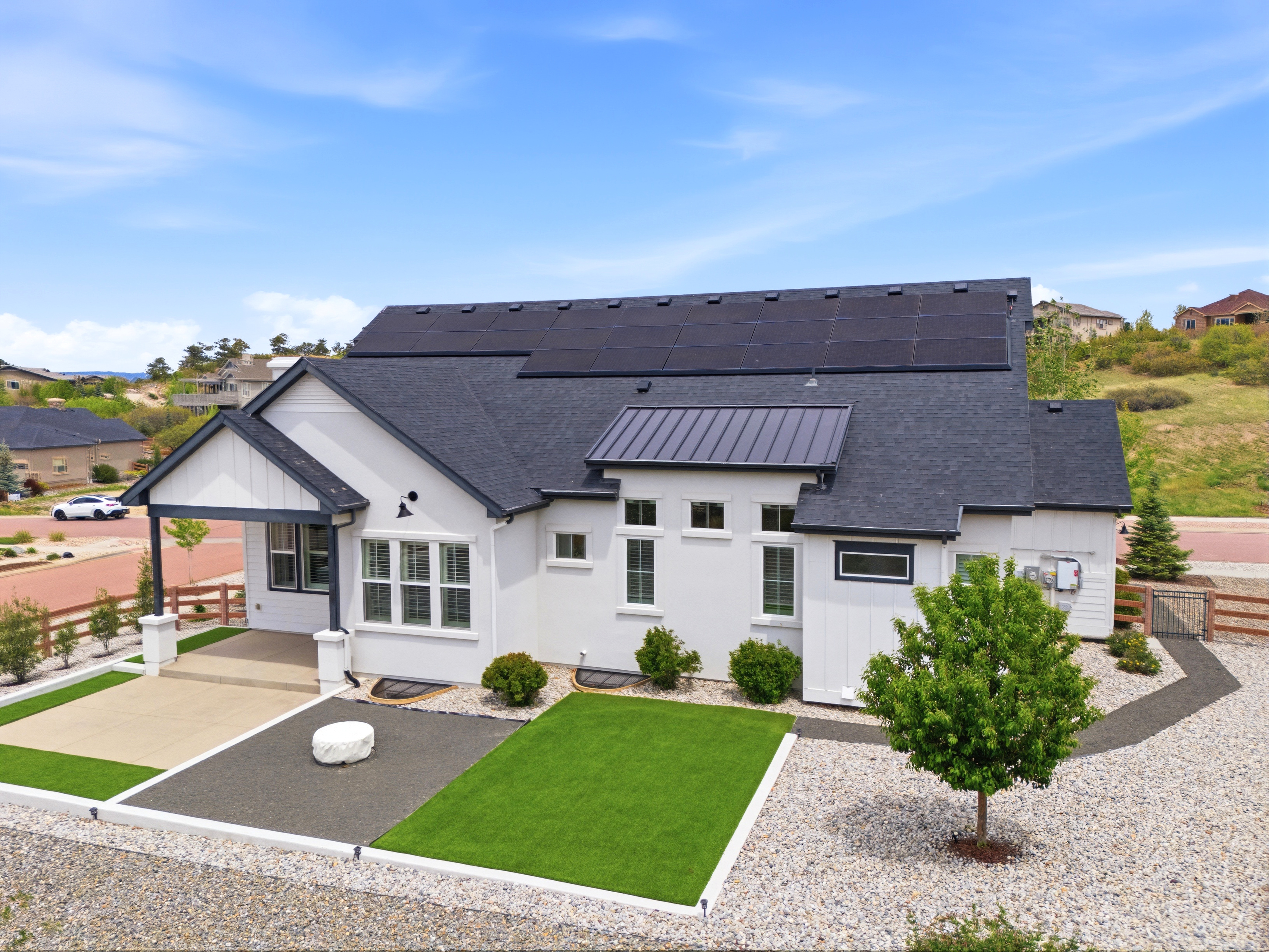 This modern farmhouse features a sleek white exterior contrasted by a dark shingle roof equipped with a large solar panel array. The property includes a low-maintenance landscape design comprising a tidy artificial turf lawn, decorative gravel, and a covered entry patio. Nestled in a scenic suburban setting, the home showcases clean lines, high-quality finishes, and a contemporary architectural aesthetic.