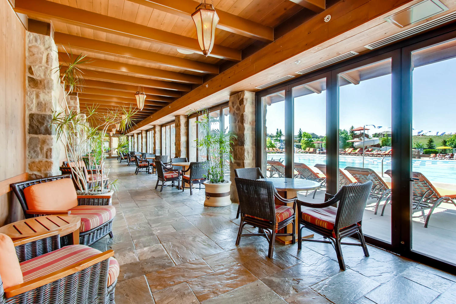 This sophisticated covered indoor lounge area features stone pillar accents, high-vaulted wood beam ceilings, and stylish wicker seating with orange-striped cushions. Large glass multi-panel doors provide a seamless transition to the outdoor pool terrace, offering a bright and relaxing space perfect for community gatherings. The design blends rustic texture with refined comfort, looking out over a bustling pool environment.