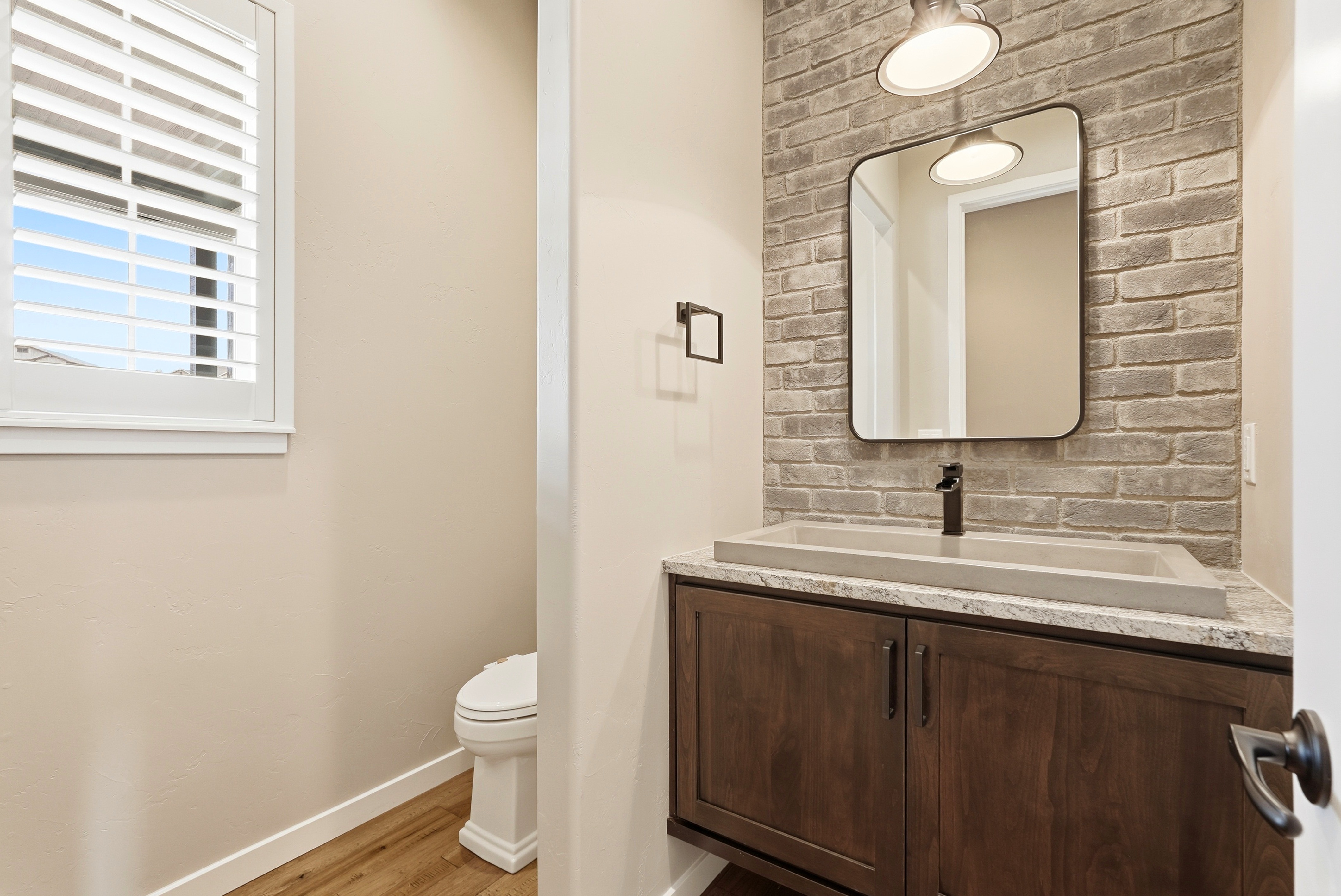 This modern guest bathroom features a unique rectangular concrete vessel sink set atop a dark wood vanity with granite-style countertops. The aesthetic is elevated by a textured gray brick accent wall, a sleek black faucet, and a contemporary mirror, complemented by warm wood flooring and clean, neutral-toned walls. The space is well-lit and thoughtfully designed with modern fixtures.