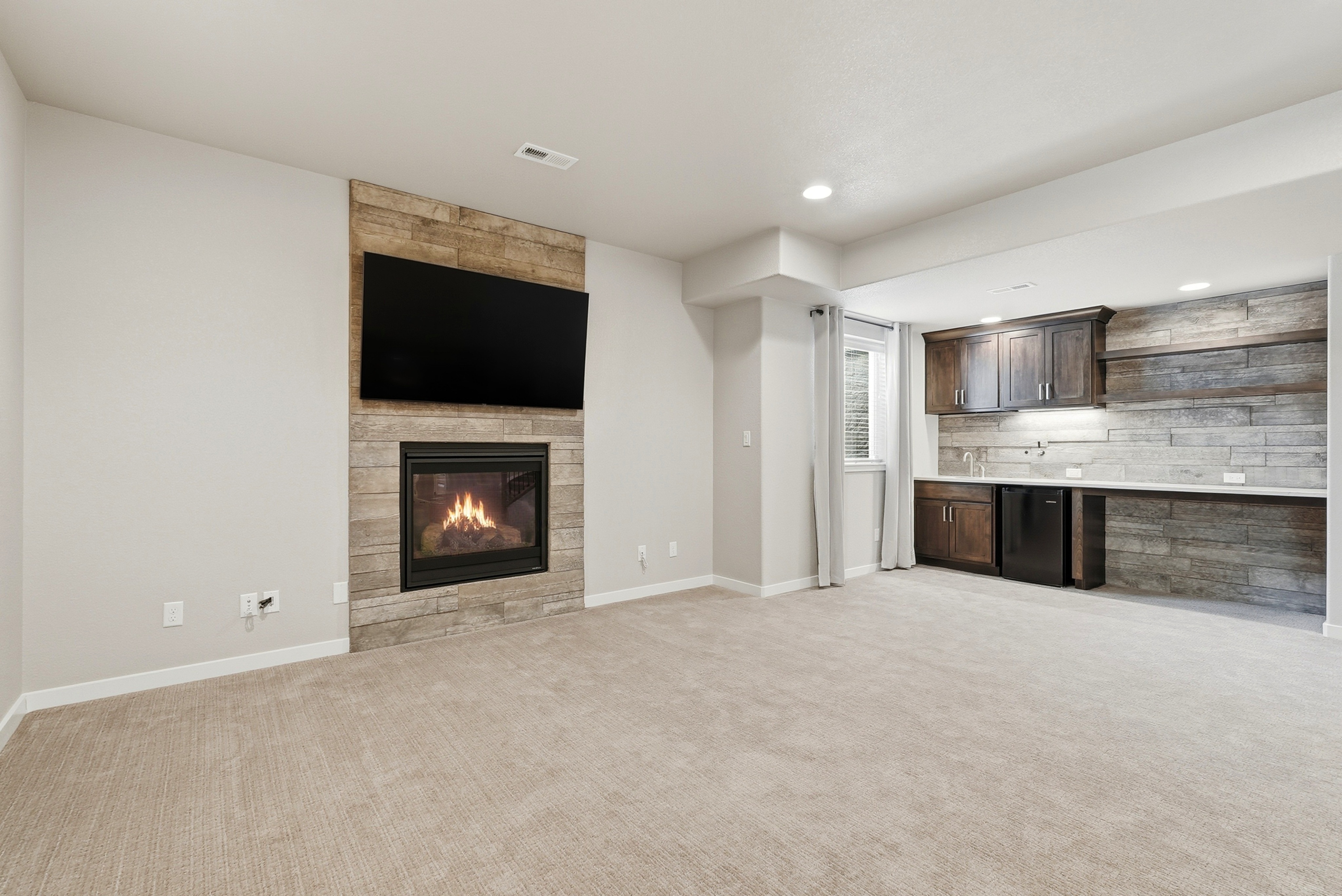 This spacious living room features a cozy fireplace with a tiled surround that doubles as a media wall for a large television. The space flows seamlessly into a wet bar area with dark wooden cabinetry, floating shelves, and a tiled backsplash, offering a functional and elegant entertainment area. The room is finished with neutral carpeting and a modern, cohesive design style.